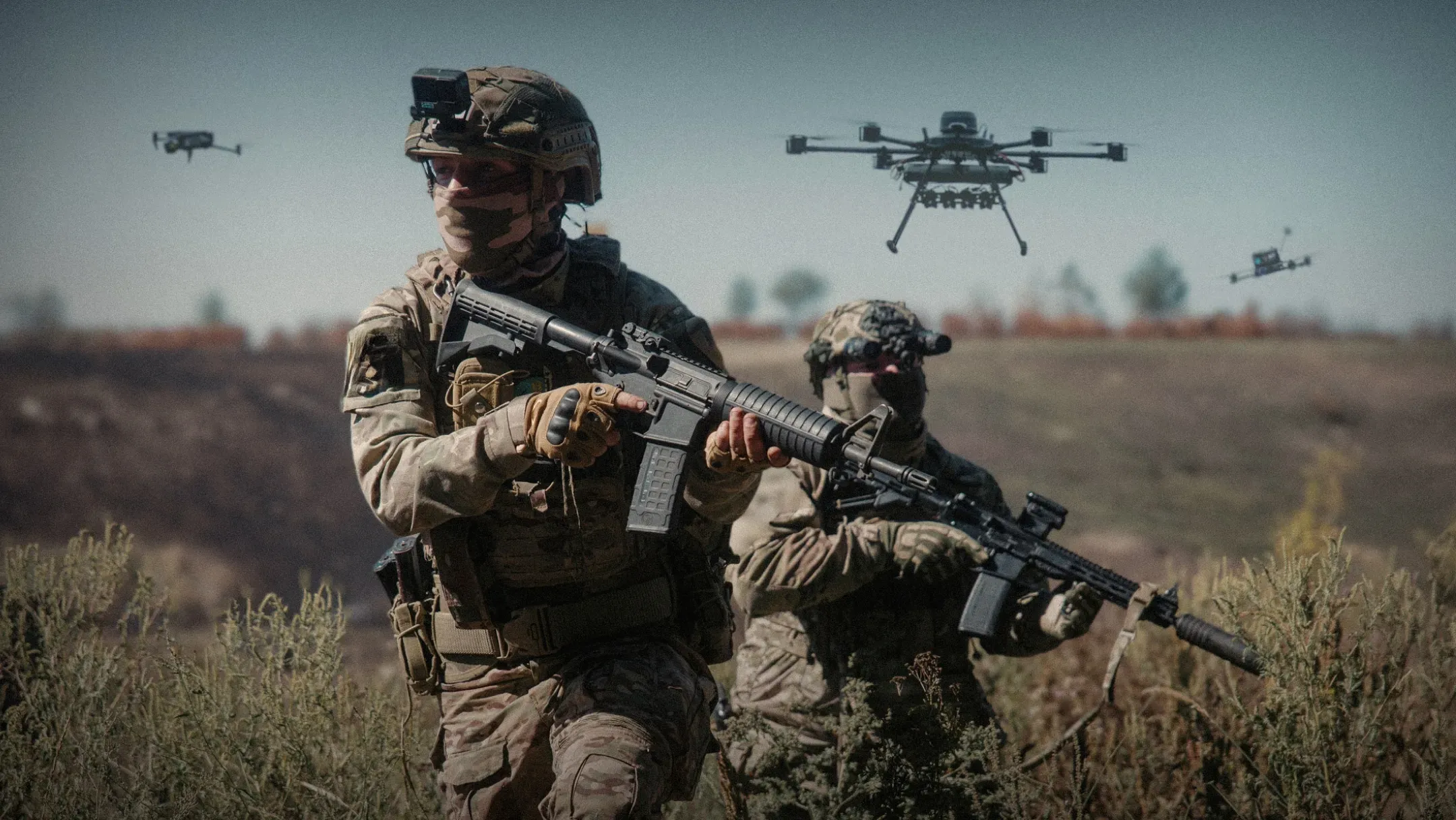 Двоє українських військових у повному бойовому спорядженні та зі штурмовими гвинтівками пересуваються полем. Їх у повітрі супроводжують три безпілотники різних типів
