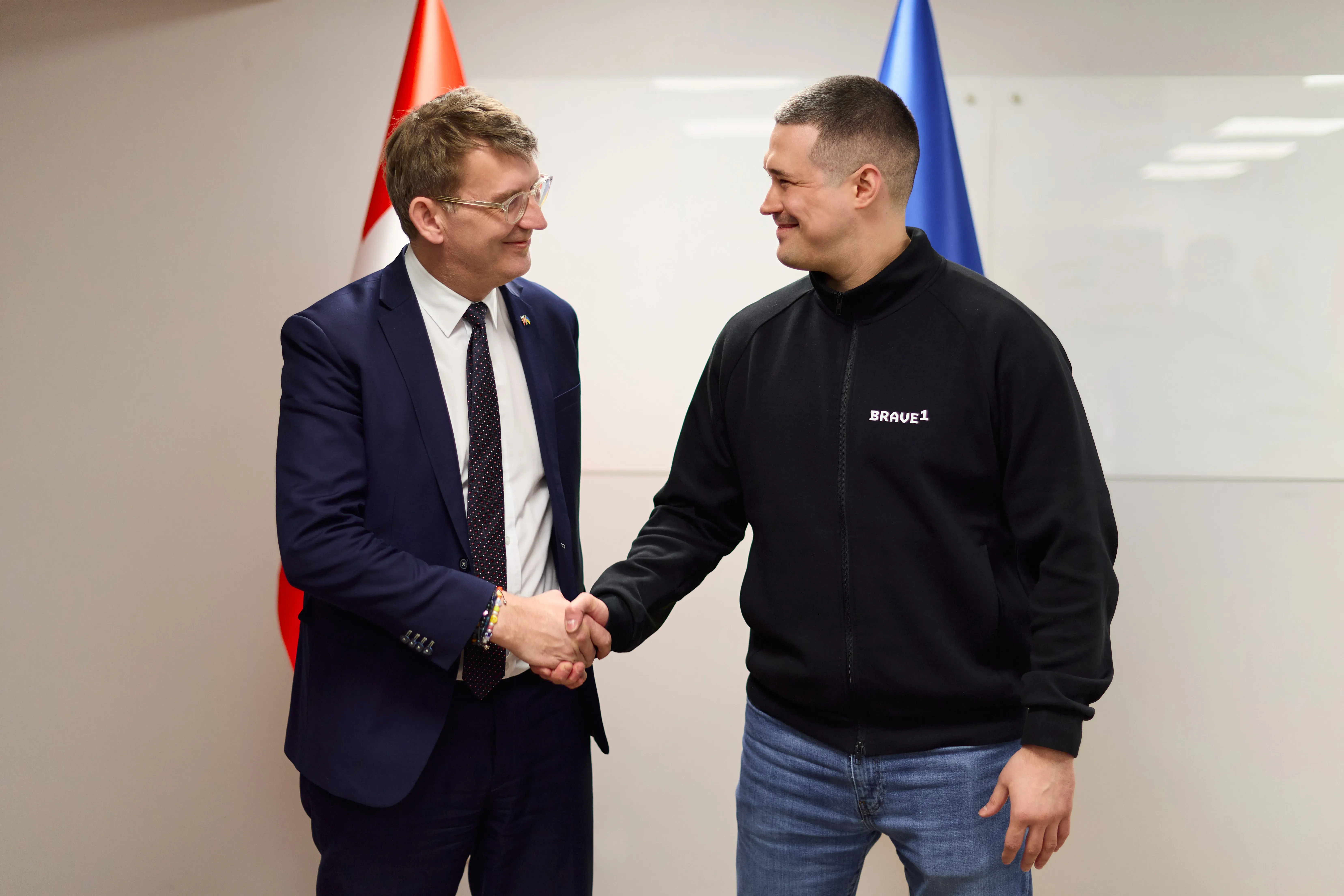 Troels Lund Poulsen (left) and Ukraine’s Minister of Defence, Mykhailo Fedorov, shake hands. Both officials look at each other and smile. The national flags of Denmark and Ukraine are partially visible in the background