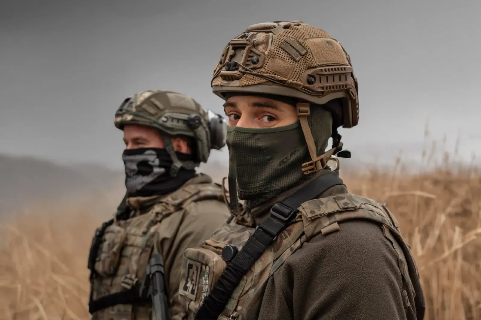 Фото двох молодих українських військовослужбовців у повному екіпіруванні на фоні відкритої місцевості. На першому плані - військовий у шоломі та захисній масці-балаклаві дивиться в камеру