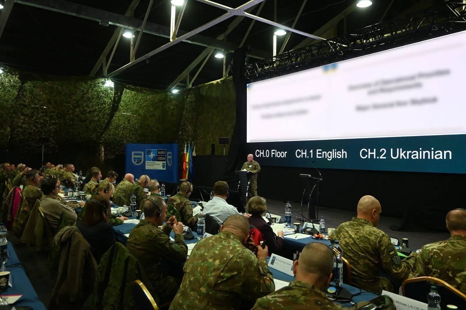 A wide view of the OFDEF conference, with a service member in a camouflage uniform speaking in front of a large screen.