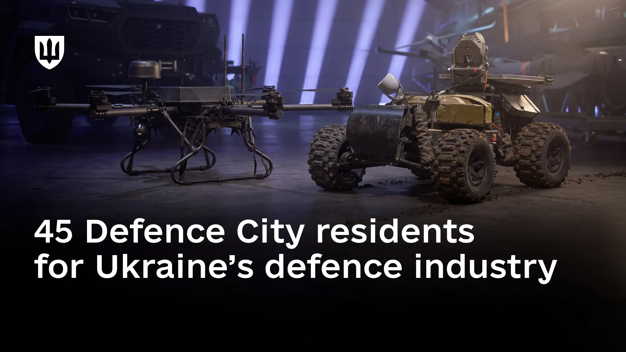 A heavy multirotor bomber drone and a four-wheeled unmanned ground vehicle stand side by side in a darkened hall, with other equipment behind them.