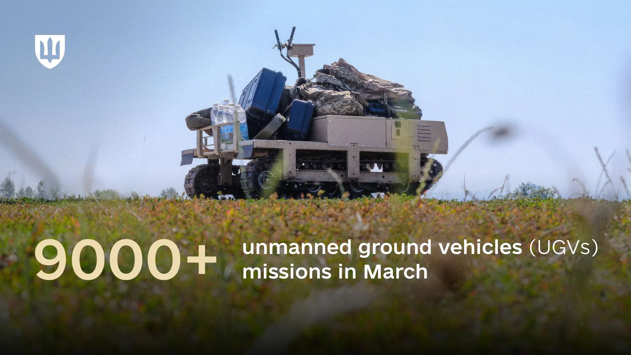 A tracked robotic ground platform traverses a field, carrying military equipment, crates, and bottles of water.