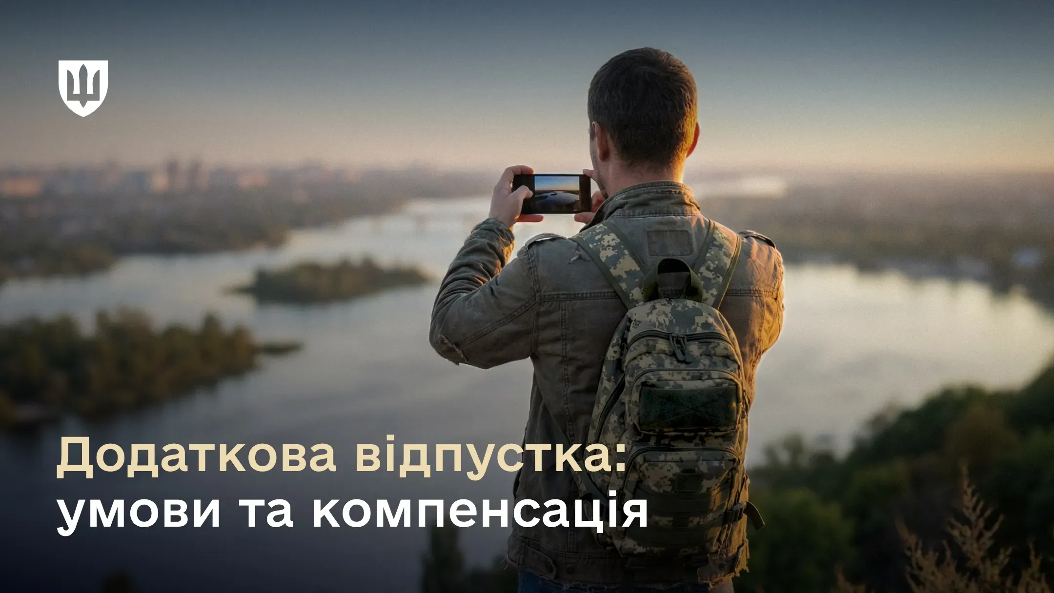 Чоловік у куртці оливкового кольору та з камуфляжним рюкзаком стоїть спиною до камери на височині. Він тримає смартфон обома руками та фотографує панораму широкої річки під час заходу сонця.