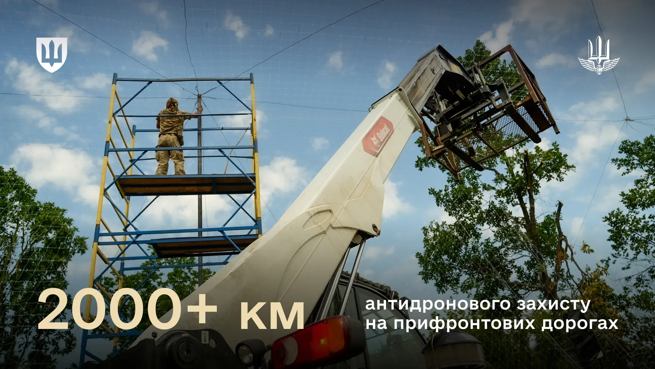 Військовослужбовець на будівельних риштуваннях монтує антидронові сітки поруч із витягнутою стрілою підіймальної спецтехніки на тлі неба та дерев