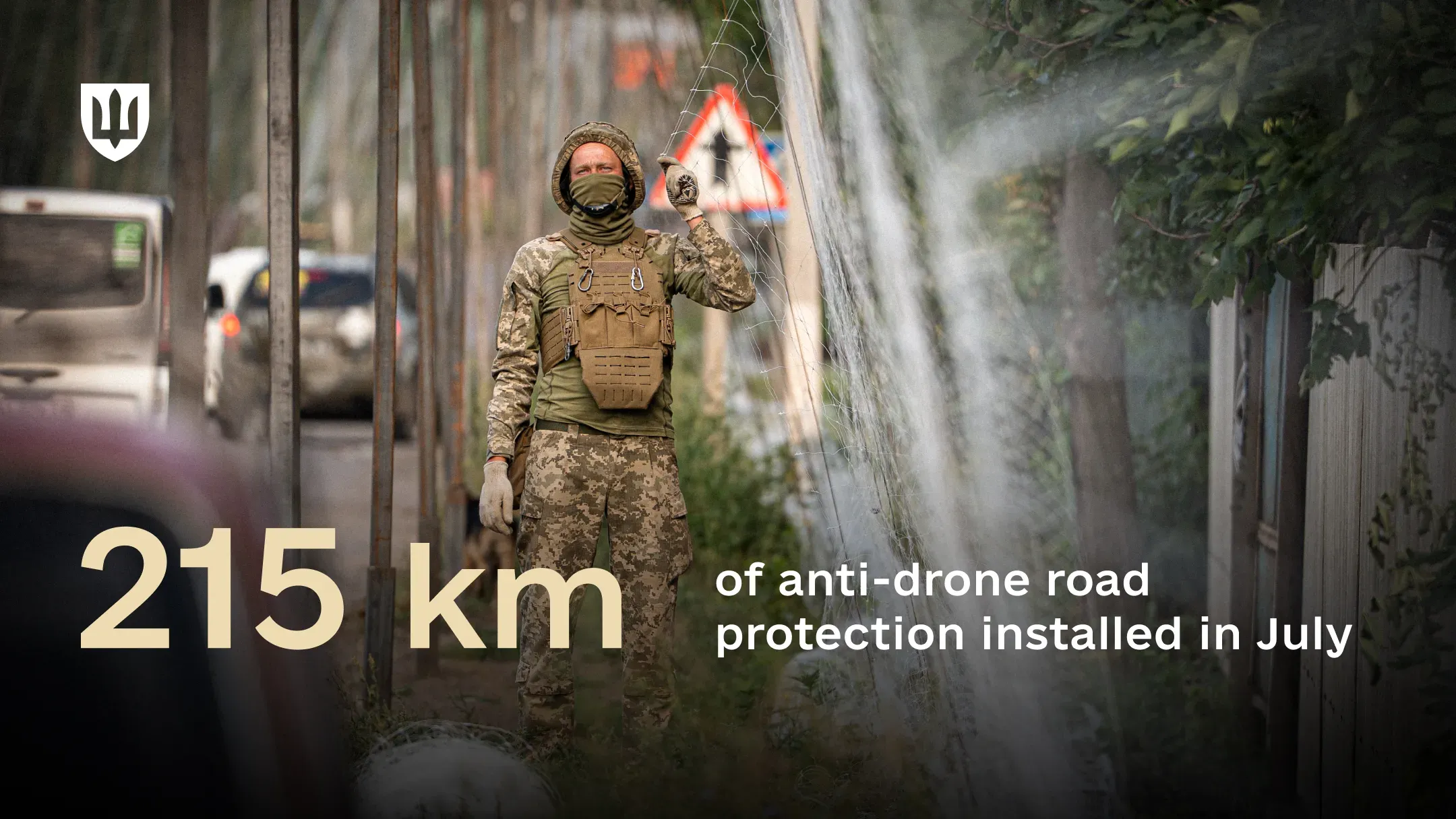 A serviceman in camouflage and a plate carrier holds up anti-drone netting strung along a roadside