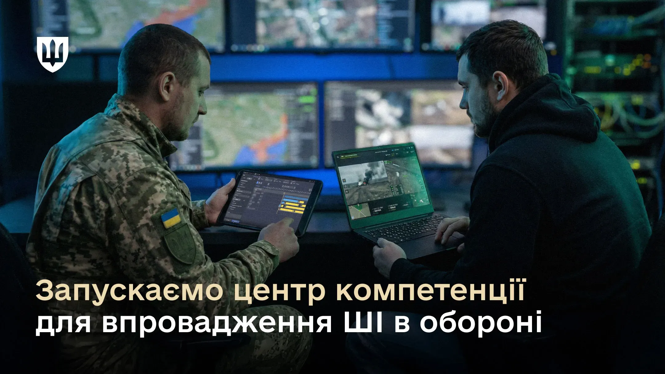 Двоє чоловіків, один у військовій формі ЗСУ, інший у цивільному одязі, працюють за ноутбуком і планшетом перед кількома екранами з картами та відео з дронів; на зображенні є напис: «Запускаємо центр компетенції для впровадження ШІ в обороні».