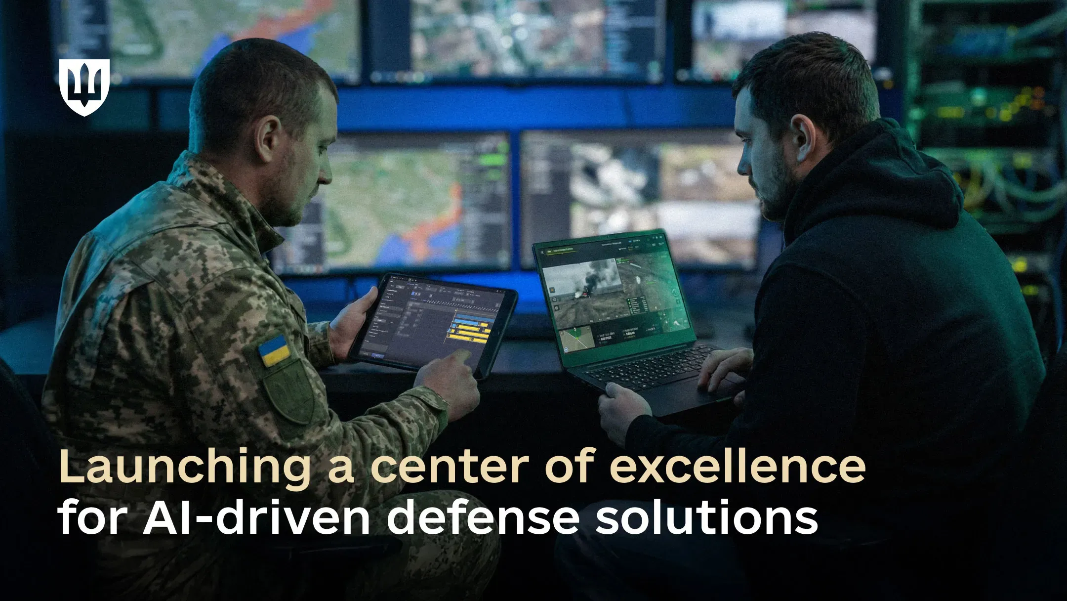 Two men—one in a Ukrainian Armed Forces uniform and the other in civilian clothing—work on a laptop and tablet in front of multiple screens displaying maps and drone.