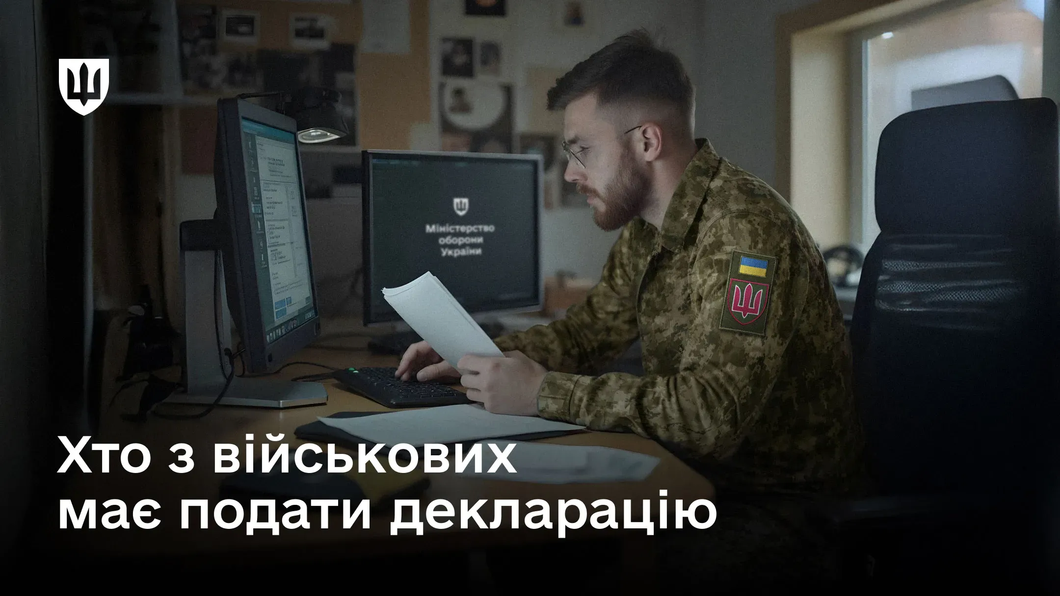 Військовий у формі Збройних Сил України сидить за робочим столом у кабінеті перед монітором та тримає в руках аркуші паперів