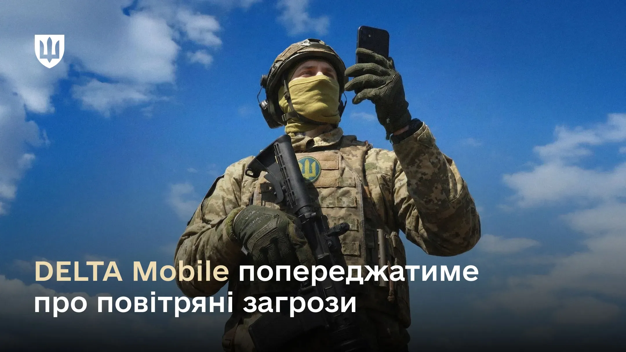 Український військовослужбовець у повному спорядженні тримає смартфон на фоні яскраво-блакитного неба