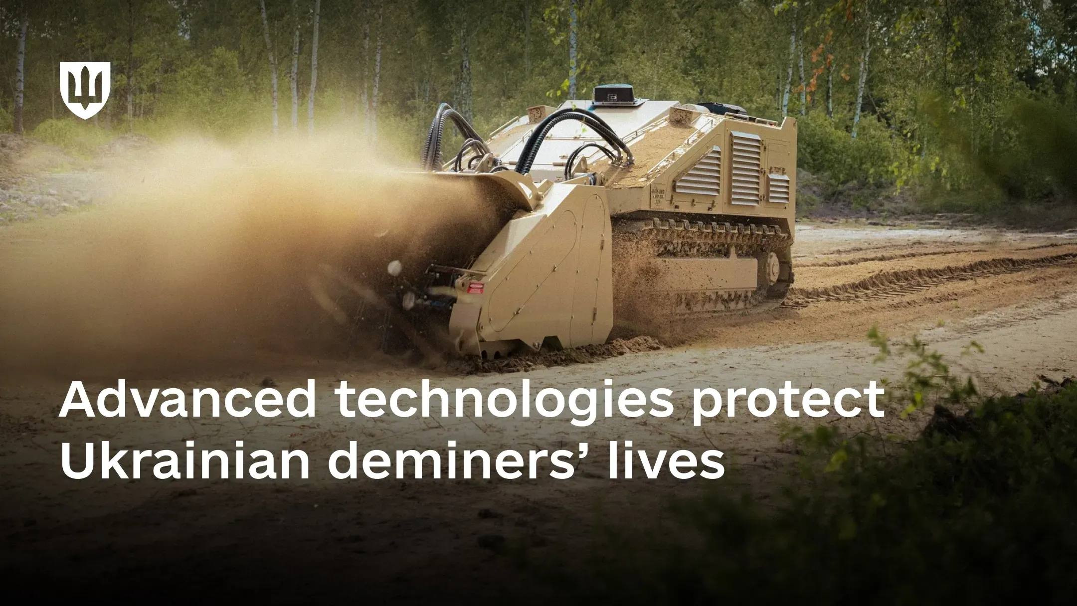 A beige tracked unmanned demining vehicle moves across a sandy area along a tree line, kicking up a large cloud of dust and sand in front of it