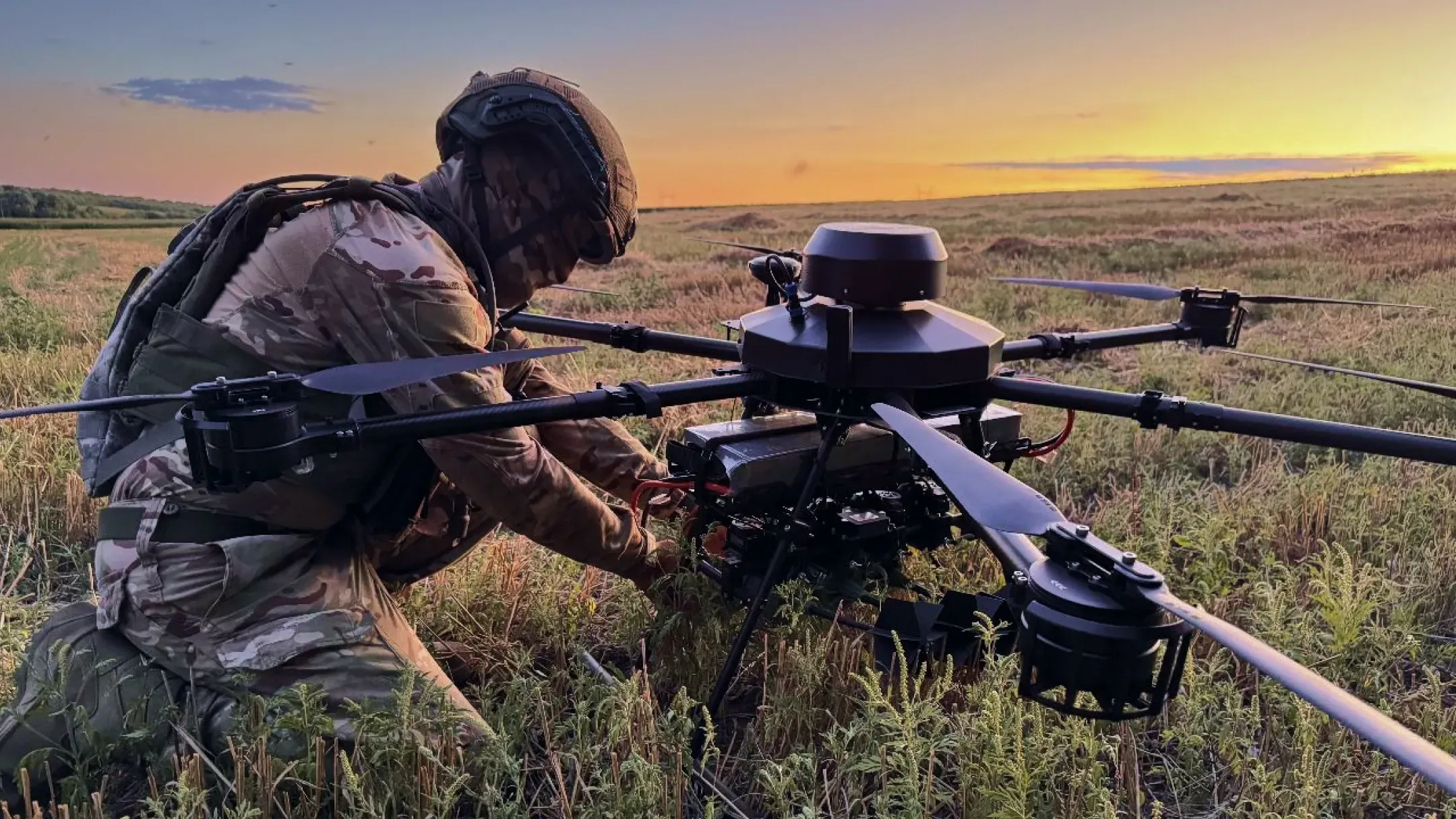A Ukrainian service member in uniform and a helmet adjusts a large eight-rotor drone in a field at sunset