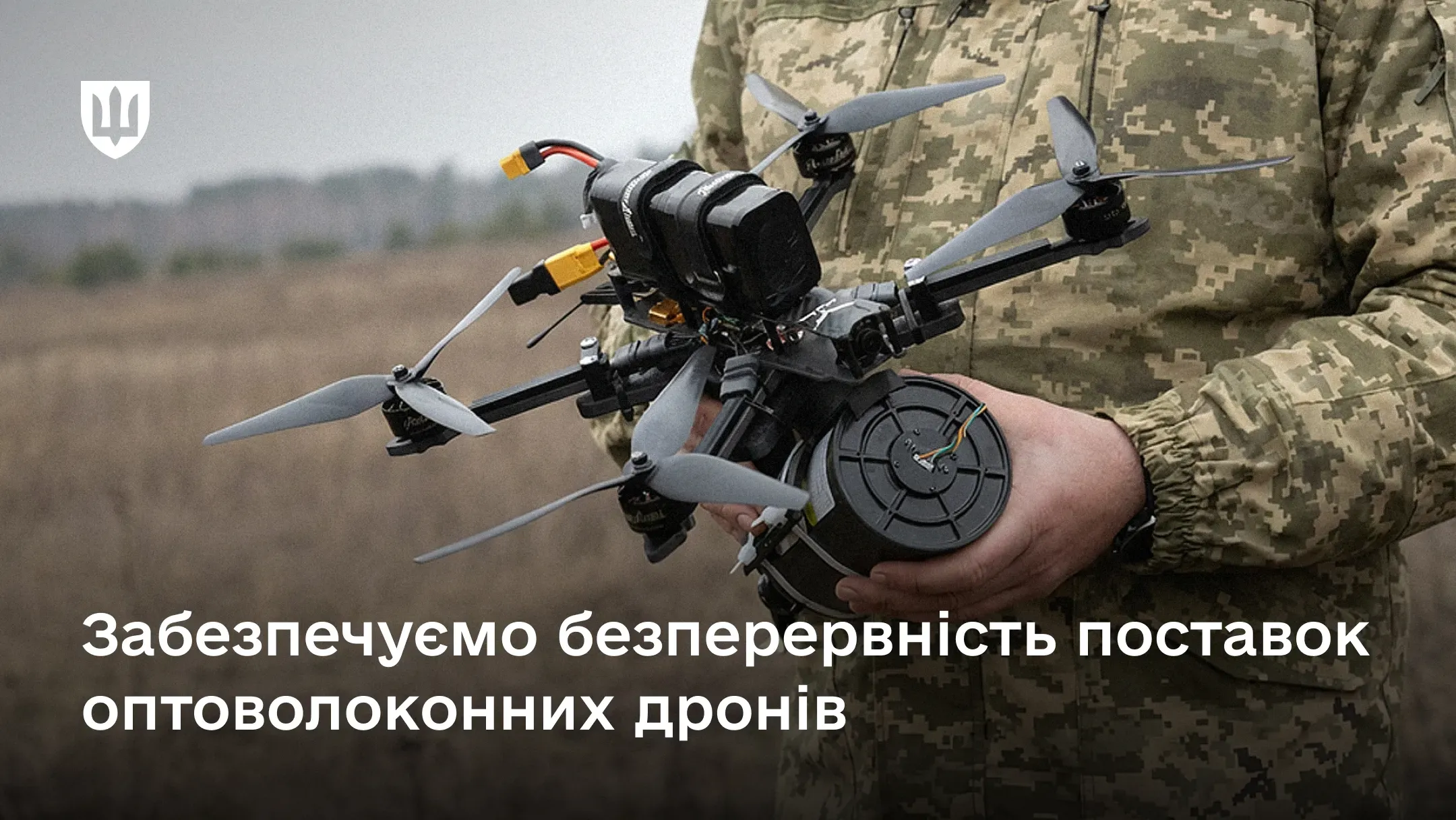 Військовослужбовець тримає в руках FPV-дрон, оснащений котушкою з оптоволокном, на фоні напису про забезпечення безперервного постачання таких безпілотників.