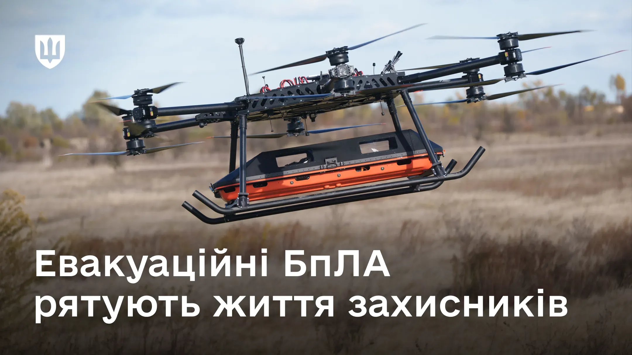 Великий багатороторний безпілотник у польоті транспортує спеціальну помаранчеву евакуаційну капсулу під фюзеляжем над відкритим полем
