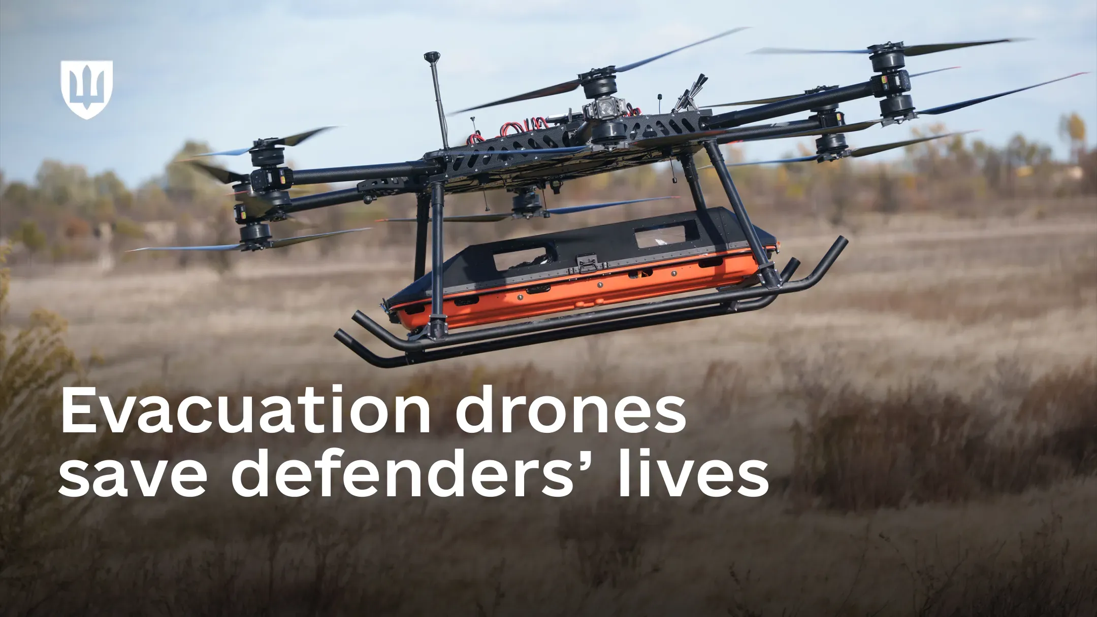 A large multirotor evacuation drone flies over an open field, carrying an orange evacuation pod beneath its fuselage. Text on image: “Evacuation UAVs save defenders’ lives”