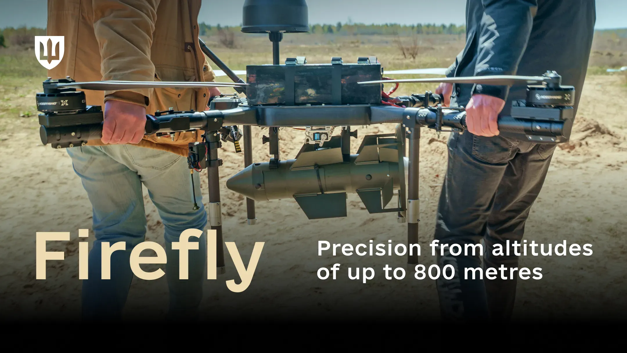 Two men carry a large multirotor bomber drone across a sandy field, with an elongated Firefly munition mounted beneath its frame