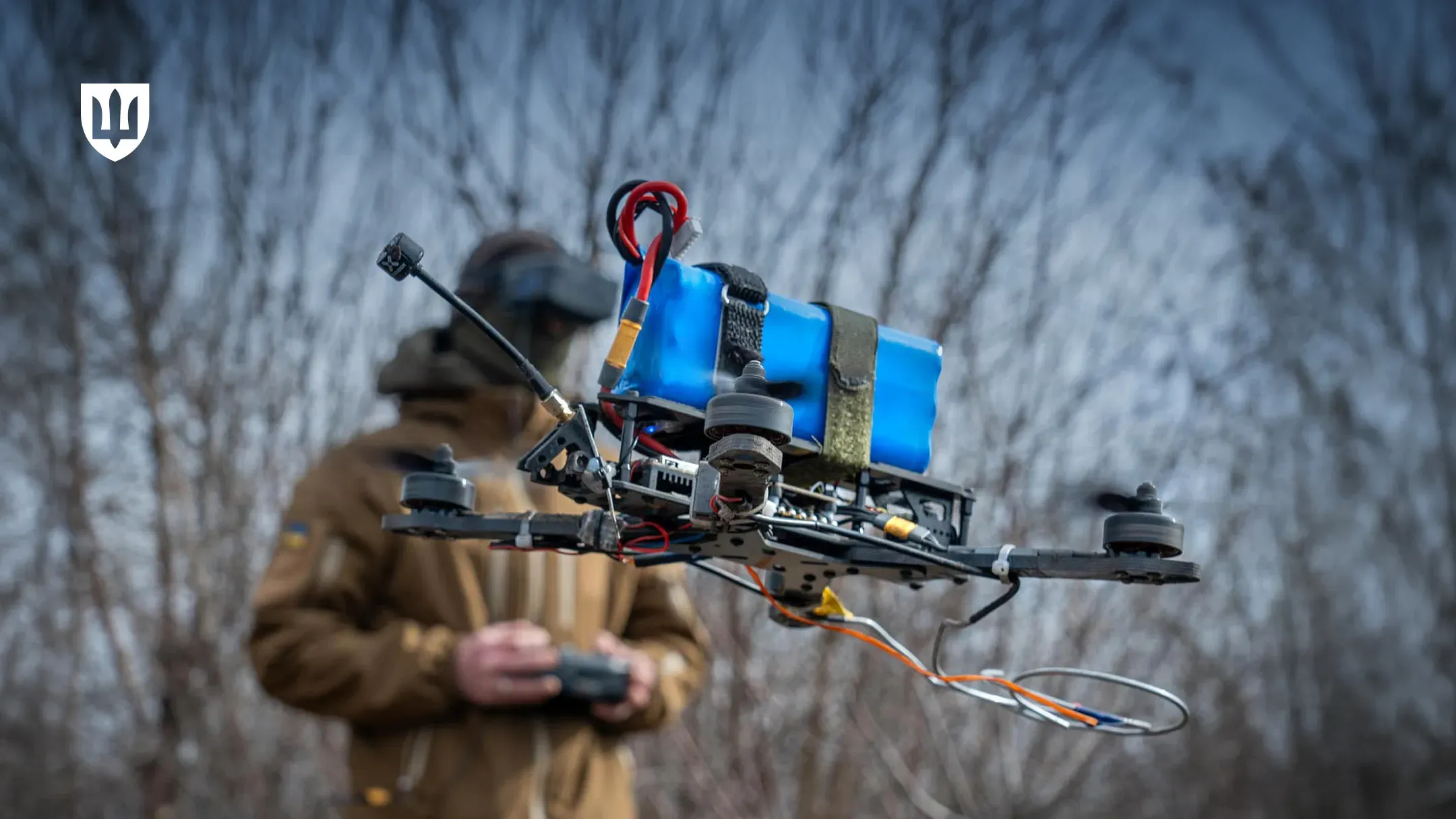 Військовий квадрокоптер FPV, що завис у повітрі. На передньому плані видно деталізовану конструкцію дрона, великий синій акумулятор, закріплений нагорі, дроти та пропелери в русі. На задньому плані стоїть український військовий у FPV-окулярах, тримаючи пульт дистанційного керування