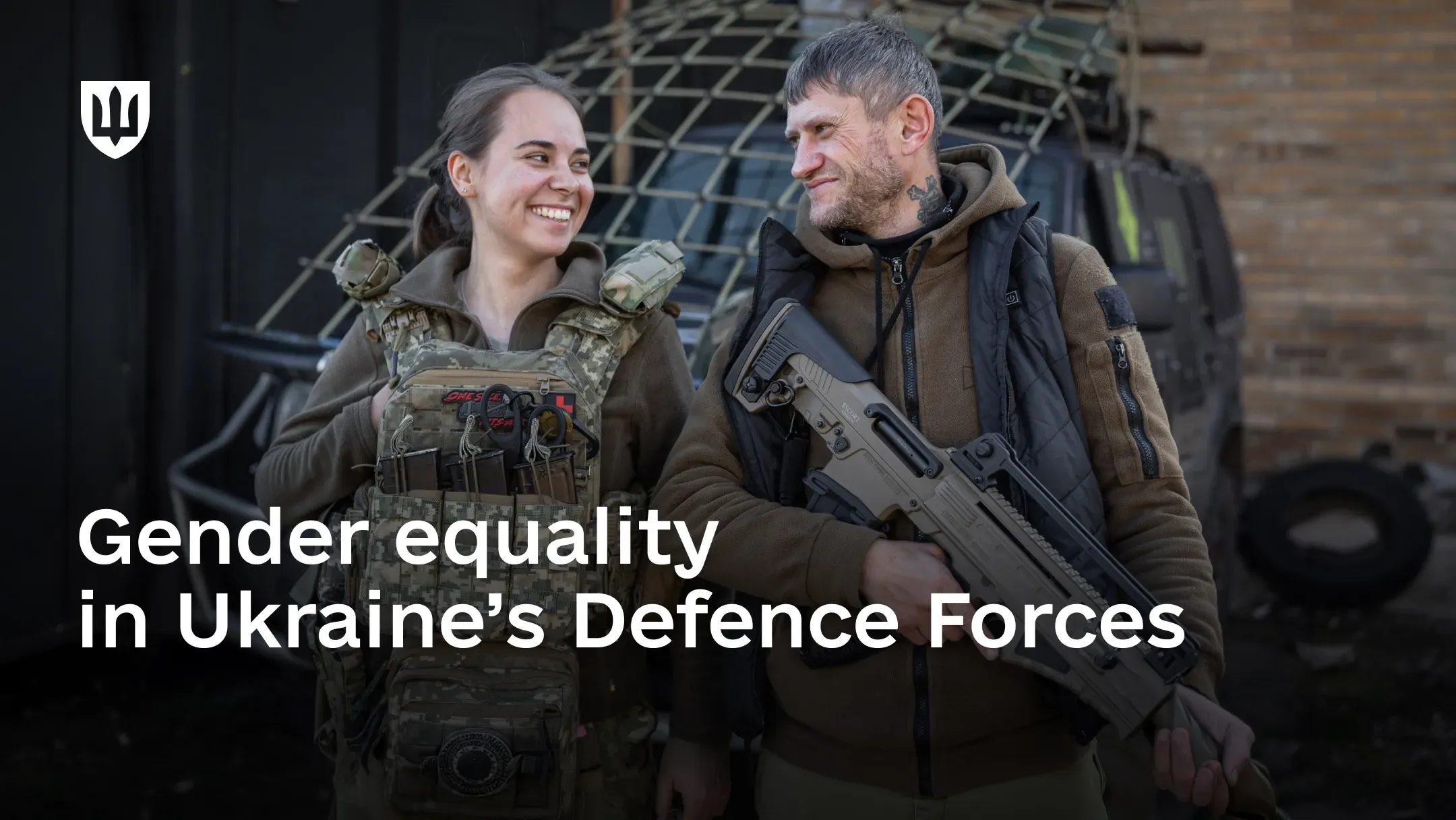 A smiling female service member in a plate carrier and a male service member holding a rifle stand in front of a vehicle covered with netting. Text on image: “Gender equality in the Defence Forces”