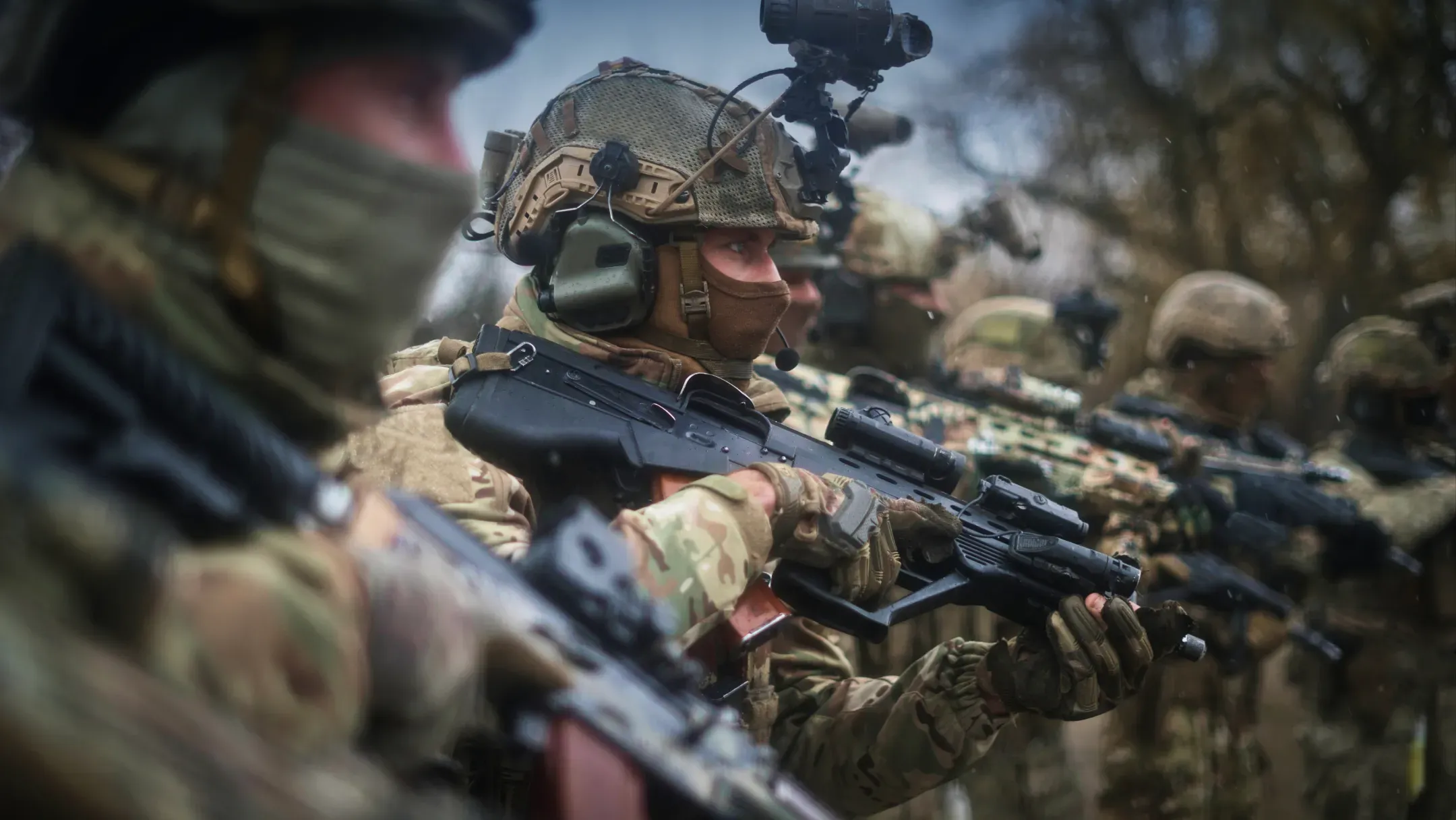 Українські військові у повному тактичному екіпіруванні та камуфляжі стоять на фоні дерев, тримаючи напоготові сучасні штурмові гвинтівки з оптичними прицілами.