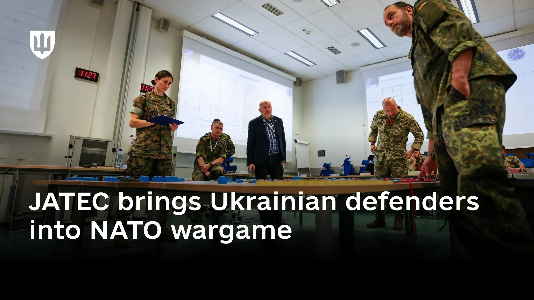 Ukrainian, German and US military personnel stand around a long table covered with coloured wargaming markers in a briefing room, with planning diagrams projected on the screens behind them