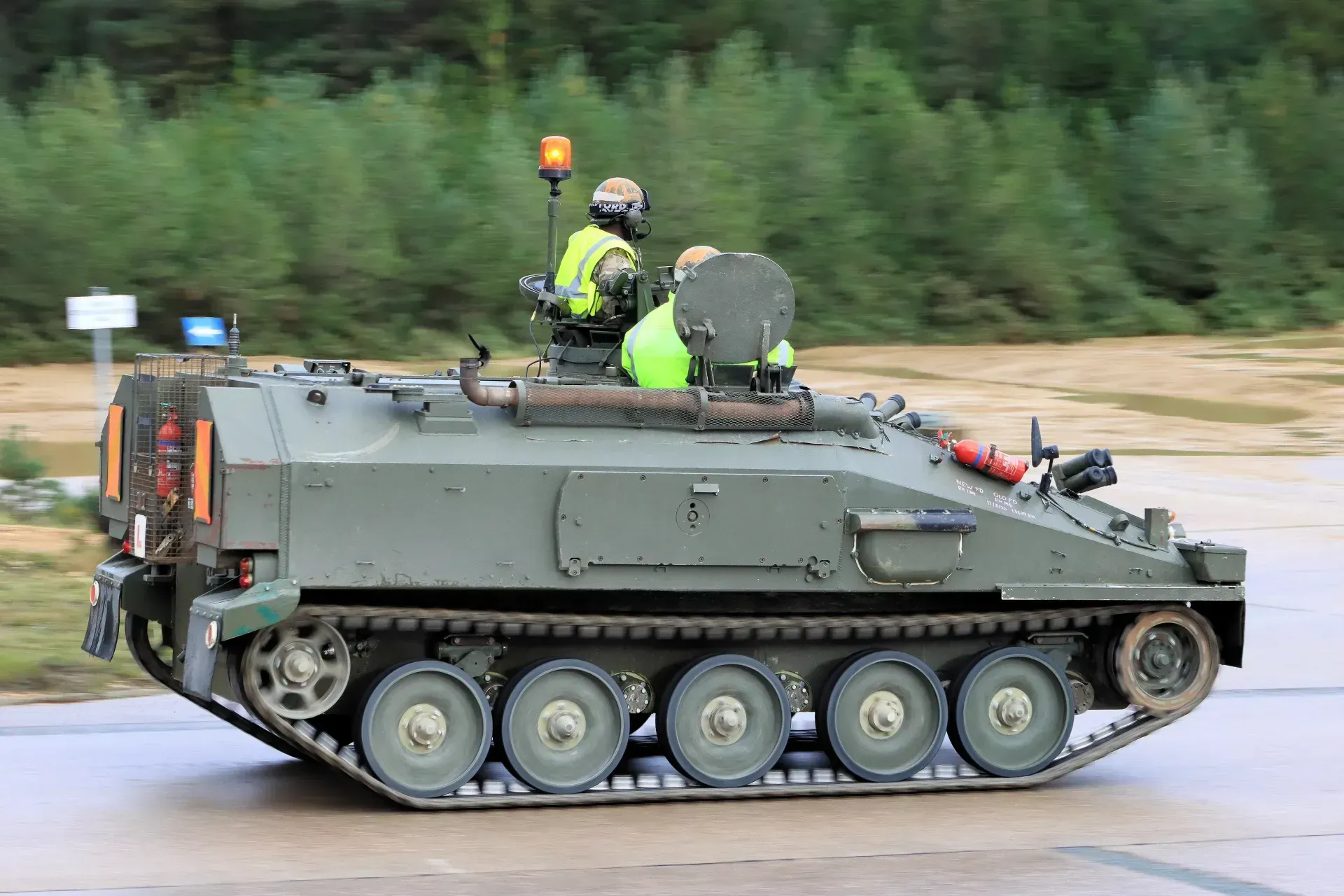 Side view of an olive-green FV103 Spartan armoured vehicle driving on a paved road. The vehicle is fitted with an orange warning beacon, and the crew are wearing high-visibility vests
