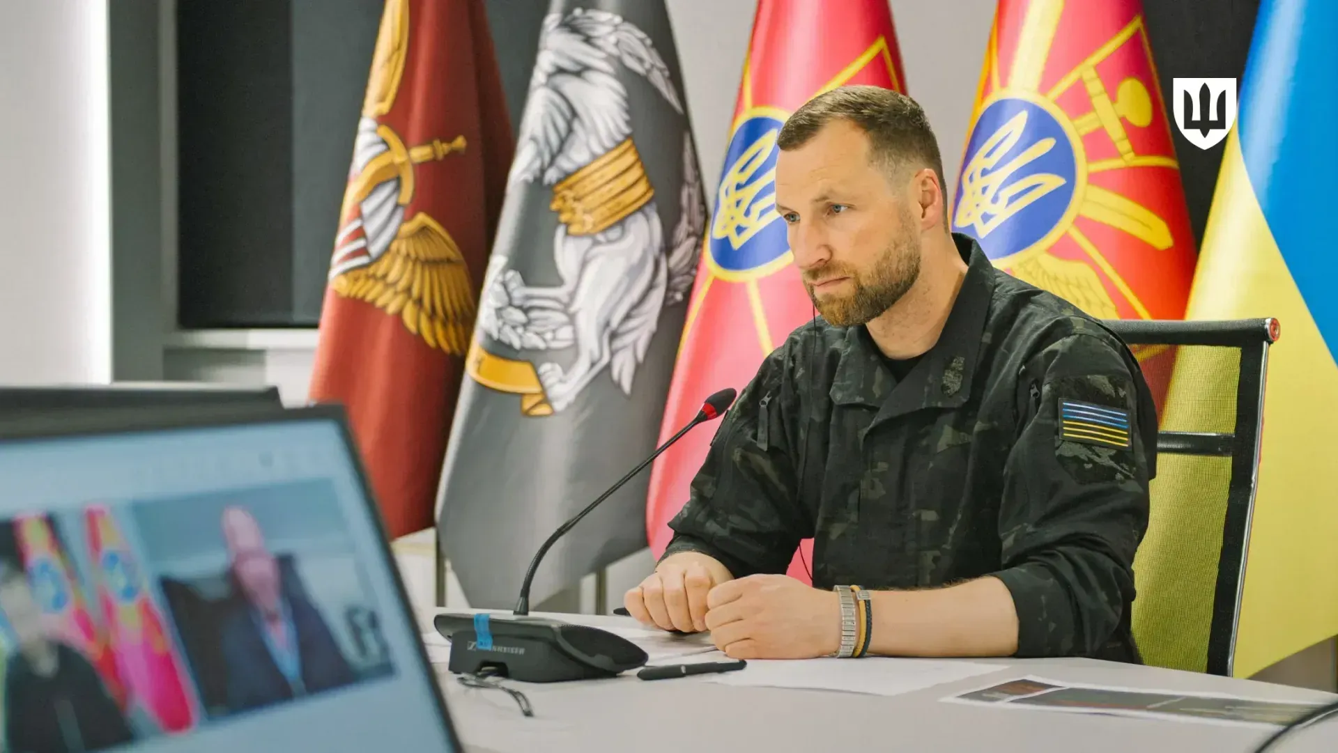 Evgeniy Khmara during a video conference, seated at a table in front of a microphone with military flags and the Ukrainian flag in the background