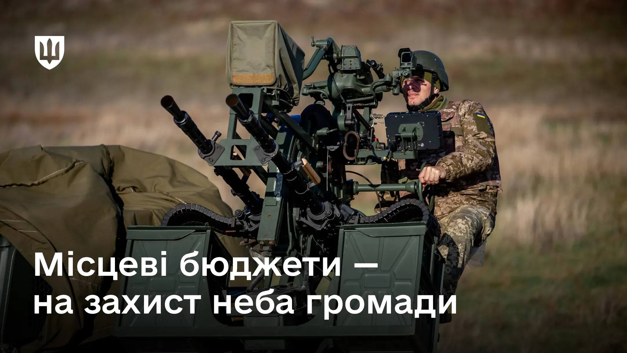 Військовослужбовець у камуфляжній формі та шоломі сидить за пультом керування спареної зенітної кулеметної установки, розташованої в полі