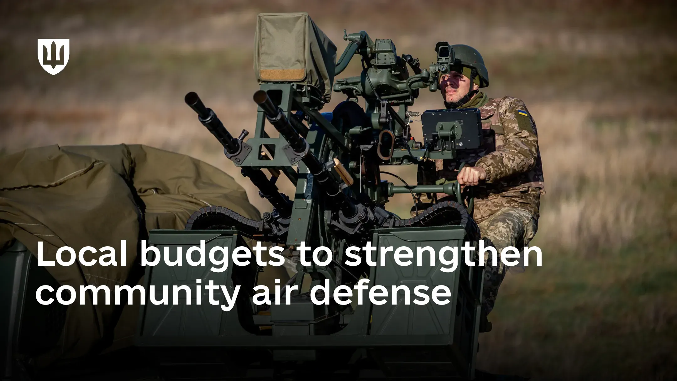 A service member in camouflage and a helmet operates a twin anti-aircraft machine-gun mount deployed in a field