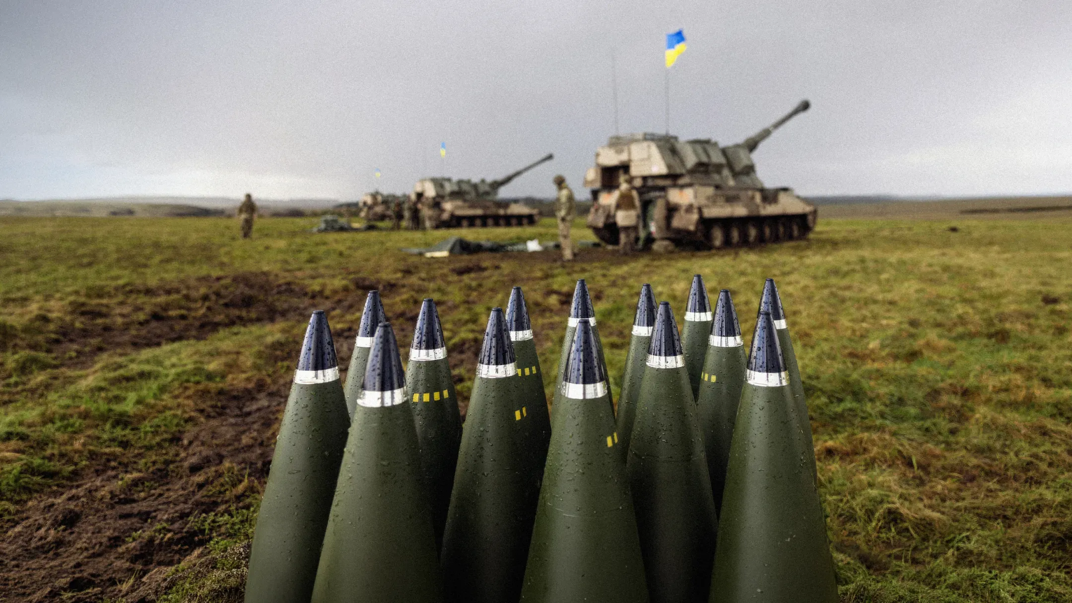 Artillery ammunition is seen in the foreground, with self-propelled artillery systems and Ukrainian flags visible in a field behind it