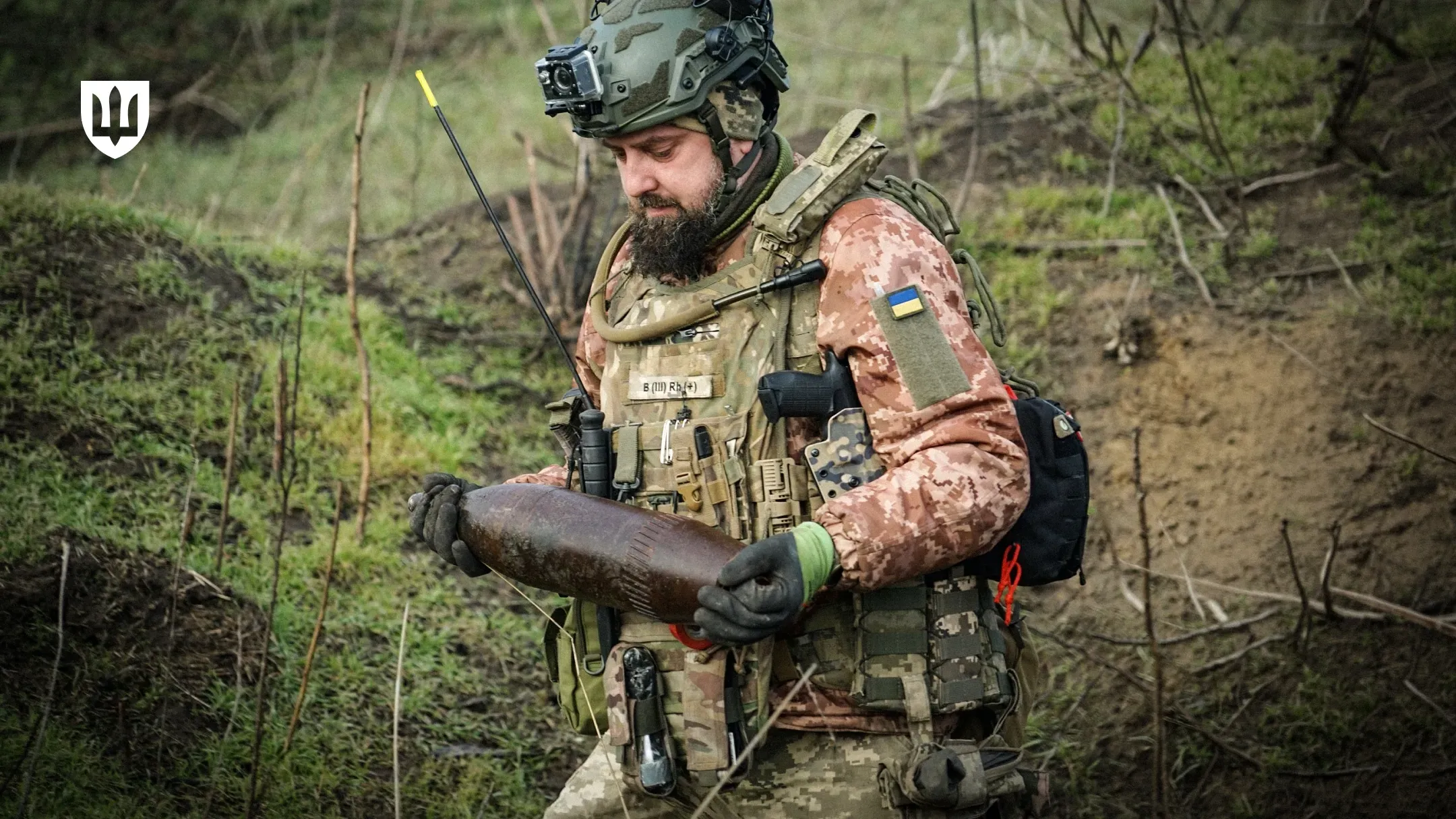 Військовослужбовець у тактичному спорядженні та шоломі тримає в руках артилерійський снаряд на відкритій місцевості