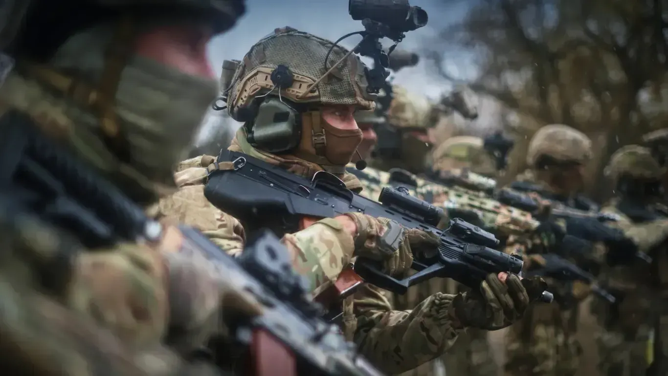 Ukrainian service members in full tactical gear and camouflage stand with trees in the background, holding modern assault rifles fitted with optical sights at the ready.