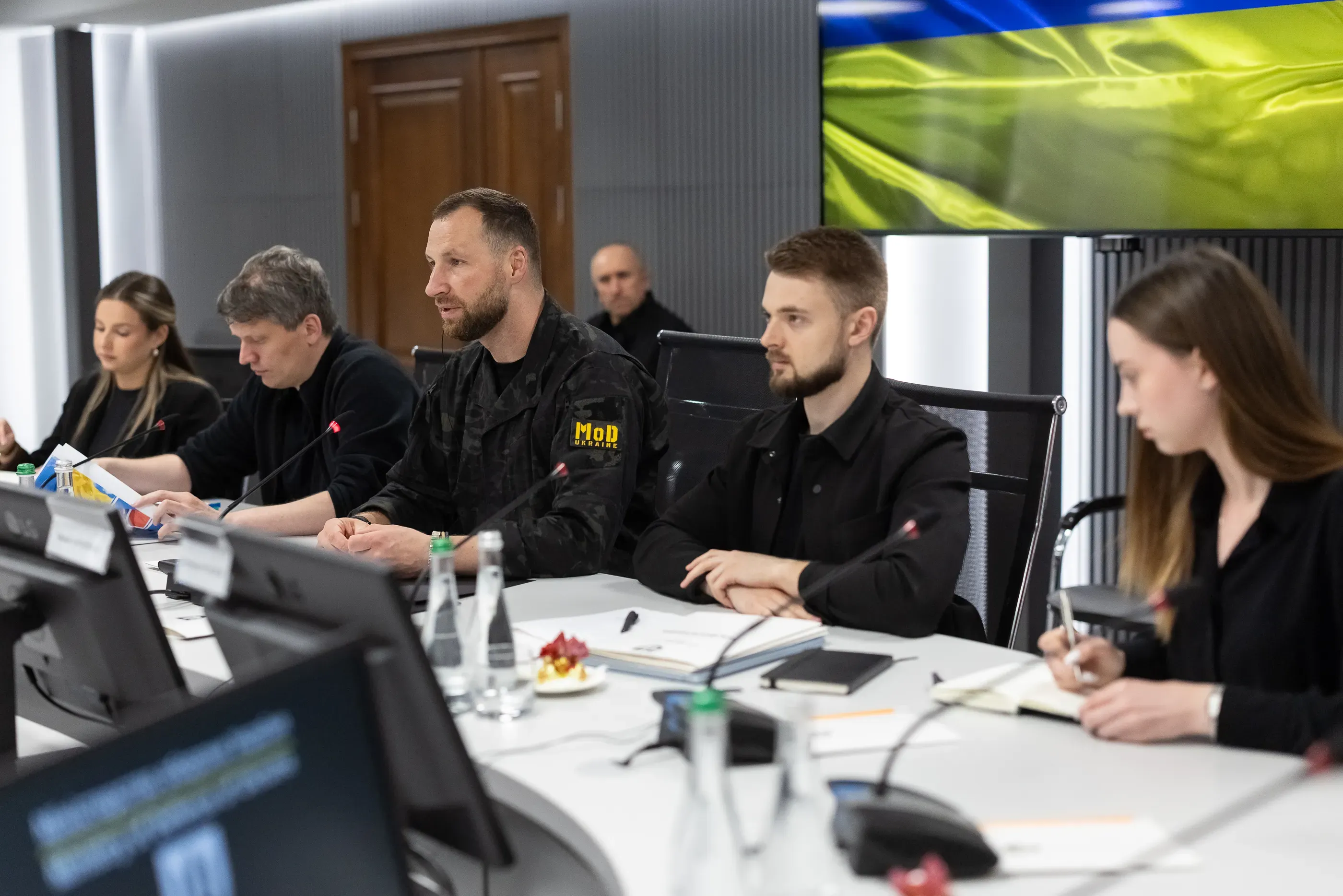 Євгеній Хмара з учасниками української делегації за столом переговорів у залі засідань на тлі екрана із зображенням прапора України