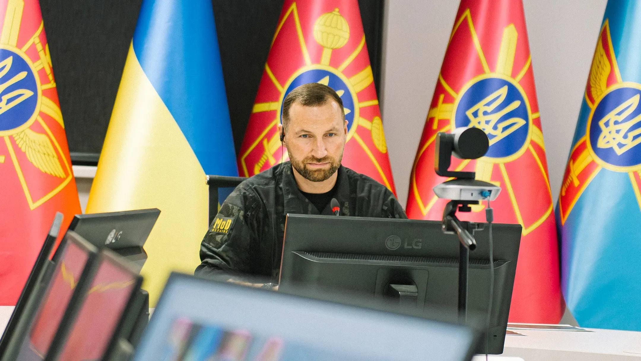Євгеній Хмара під час телефонної розмови за робочим столом перед монітором і камерою, на тлі державного прапора України та військових стягів