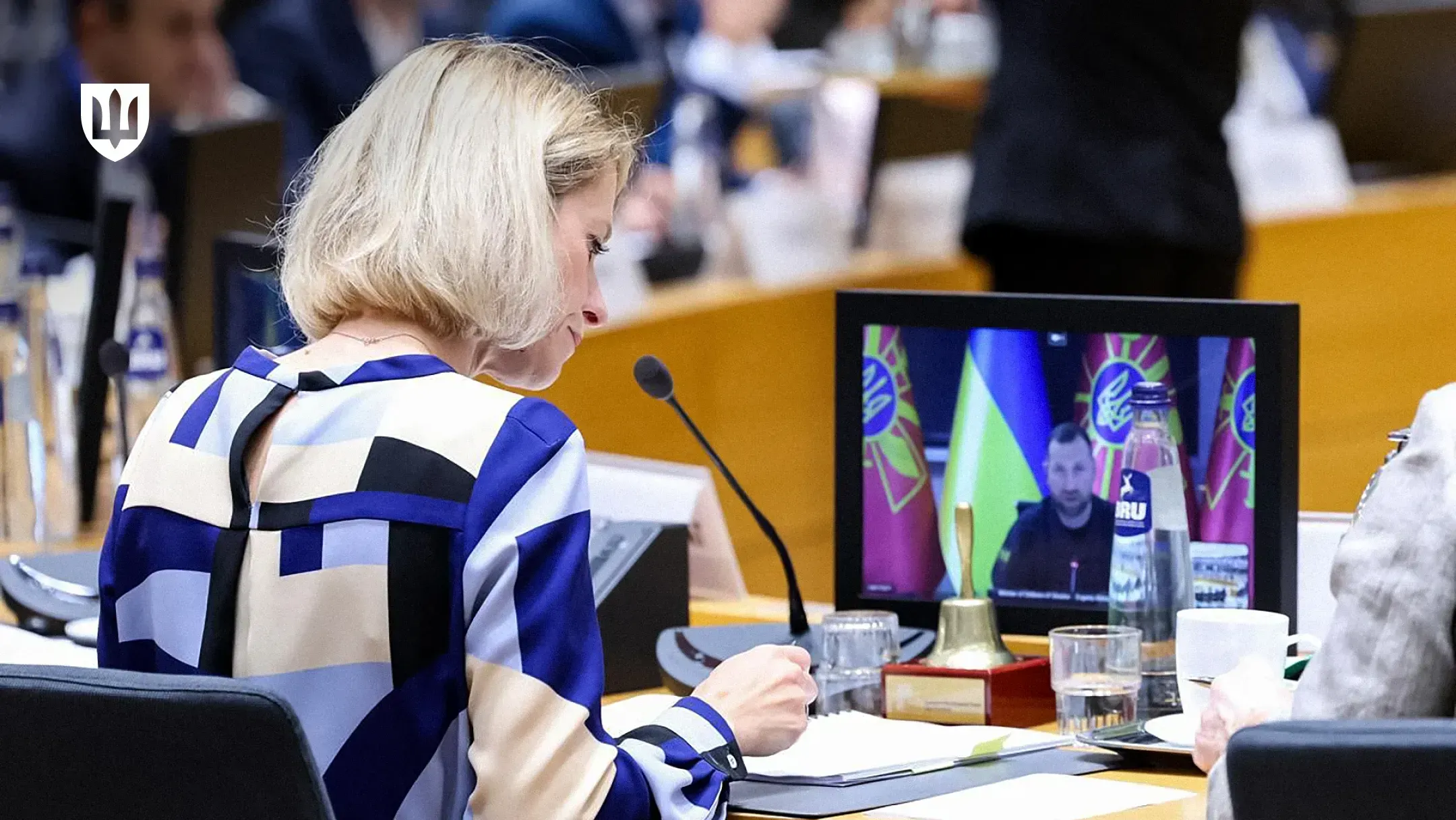 Kaja Kallas taking notes during an EU Council meeting, with Evgeniy Khmara appearing by video link on a monitor in front of her