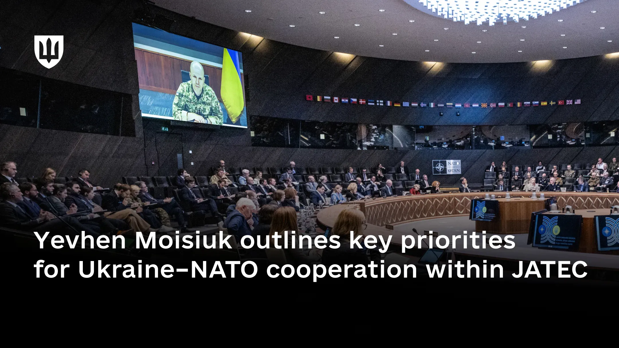 A large circular conference hall during a meeting. On a large screen, Lieutenant General Yevhen Moisiuk, Deputy Minister of Defence of Ukraine, appears in pixel-pattern uniform while addressing the participants.