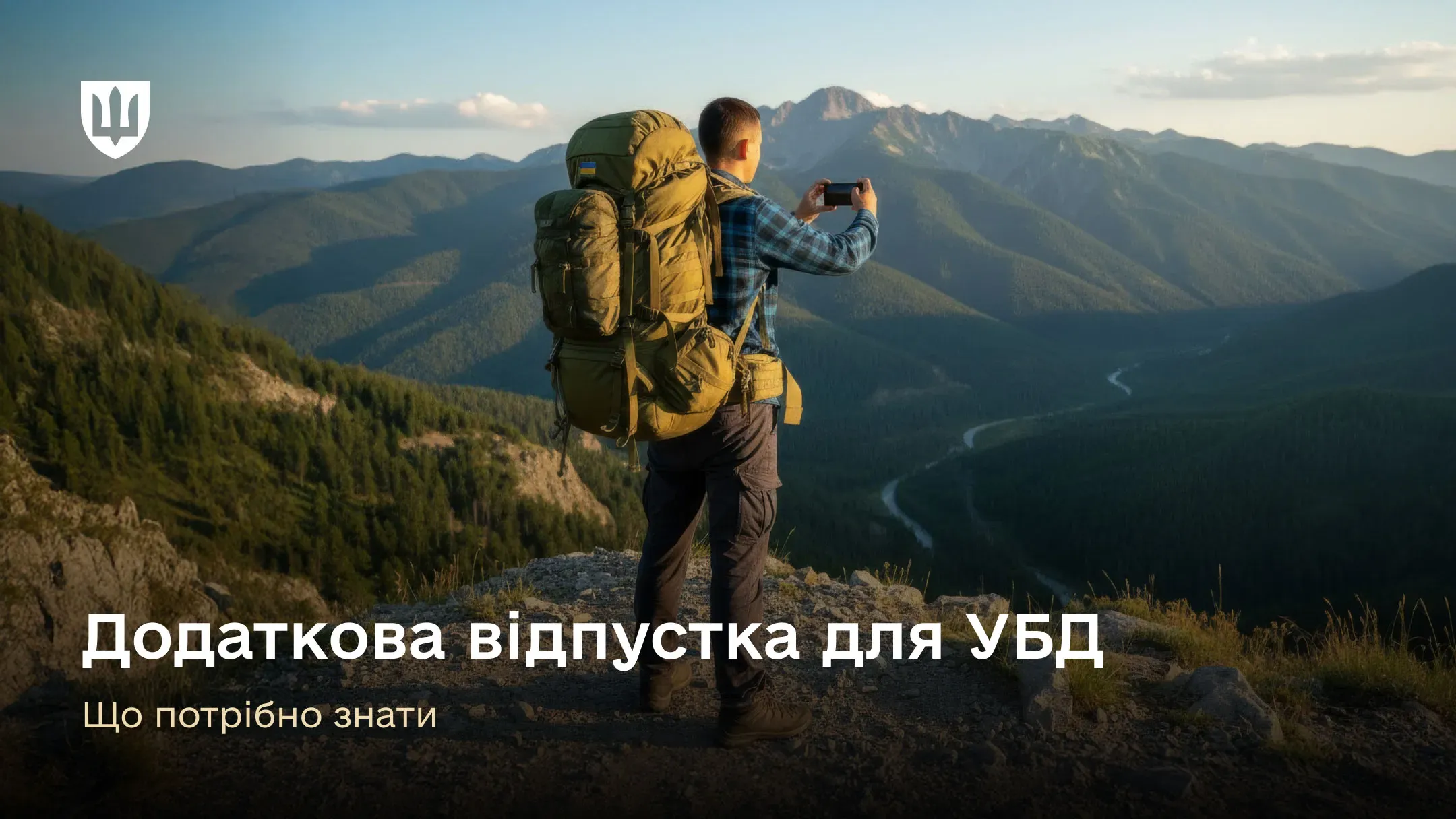 Чоловік у туристичному спорядженні з великим наплічником з прапором України стоїть на вершині гори, дивлячись на мальовничий гірський пейзаж, та фотографує його на смартфон. 