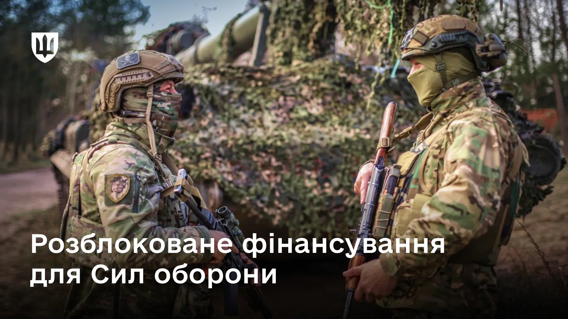 Двоє українських військовослужбовців спілкуються біля замаскованої бронетехніки в лісовій місцевості