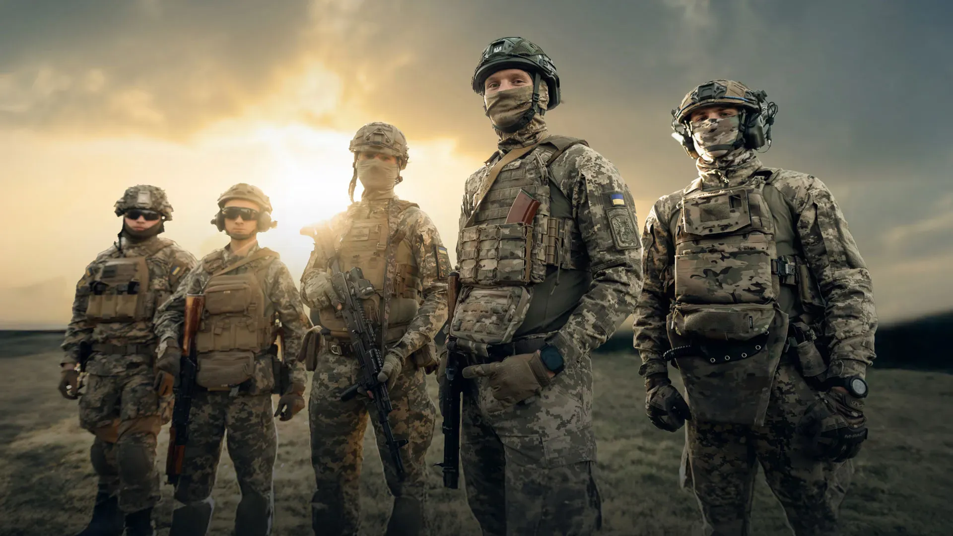 Five Ukrainian service members in full modern gear stand together against a cloudy sky lit by the setting sun