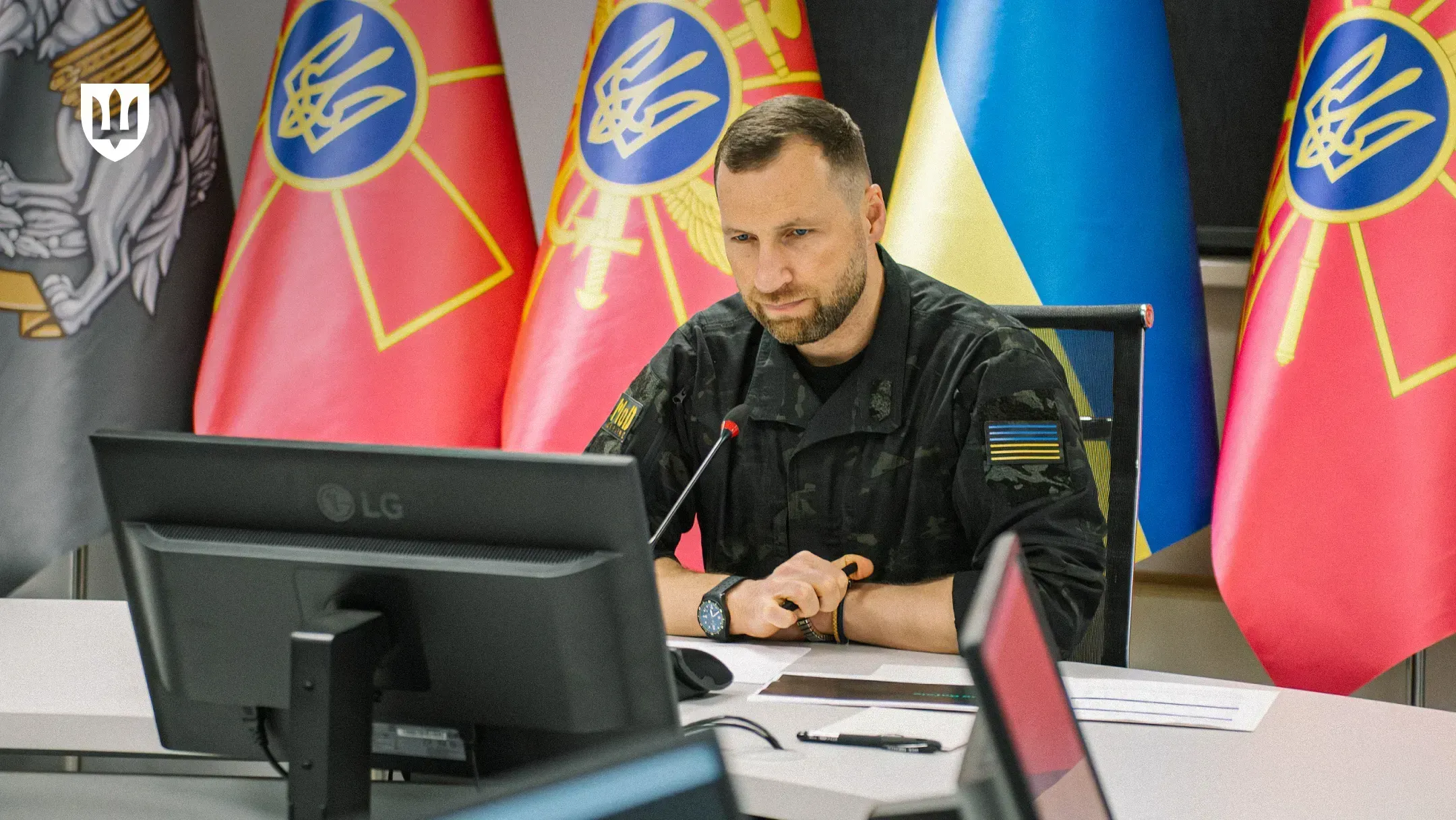 Evgeniy Khmara seated at a table in front of a monitor and microphone during an online meeting, with the national flag of Ukraine and Ministry of Defence flags in the background