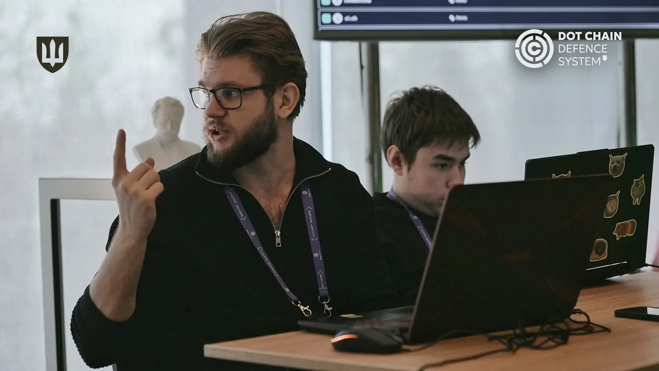 Чоловік з бородою та в окулярах, одягнений у чорний светр, сидить за робочим столом із ноутбуком і емоційно щось пояснює, піднявши вказівний палець вгору. Поруч із ним інший спеціаліст зосереджено працює за комп'ютером