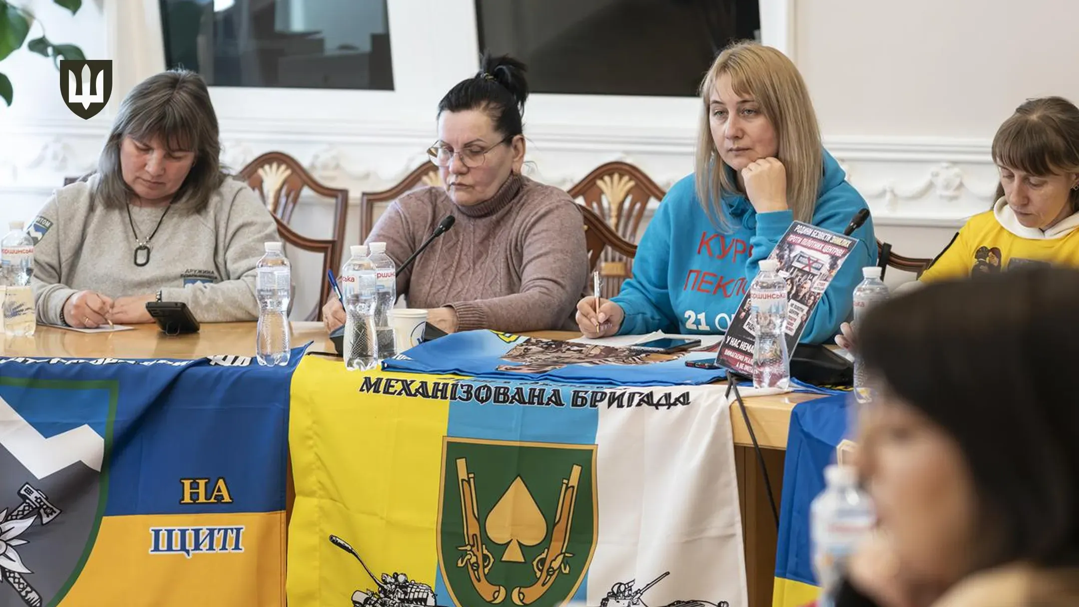 Жінки сидять за столом під час офіційної зустрічі або пресконференції. Перед ними розстелені прапори та шеврони українських військових підрозділів. На столі стоять пляшки води та мікрофони.