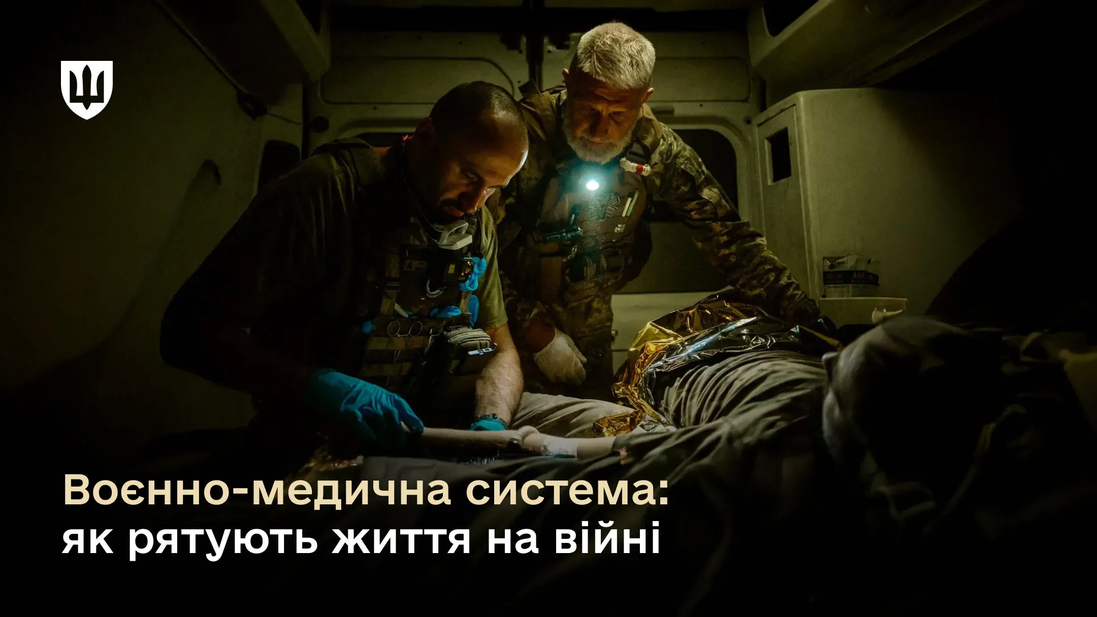 Художнє фото двох українських бойових медиків у повному спорядженні з налобними ліхтариками, які зосереджено надають допомогу пораненому в напівтемряві всередині евакуаційного автомобіля