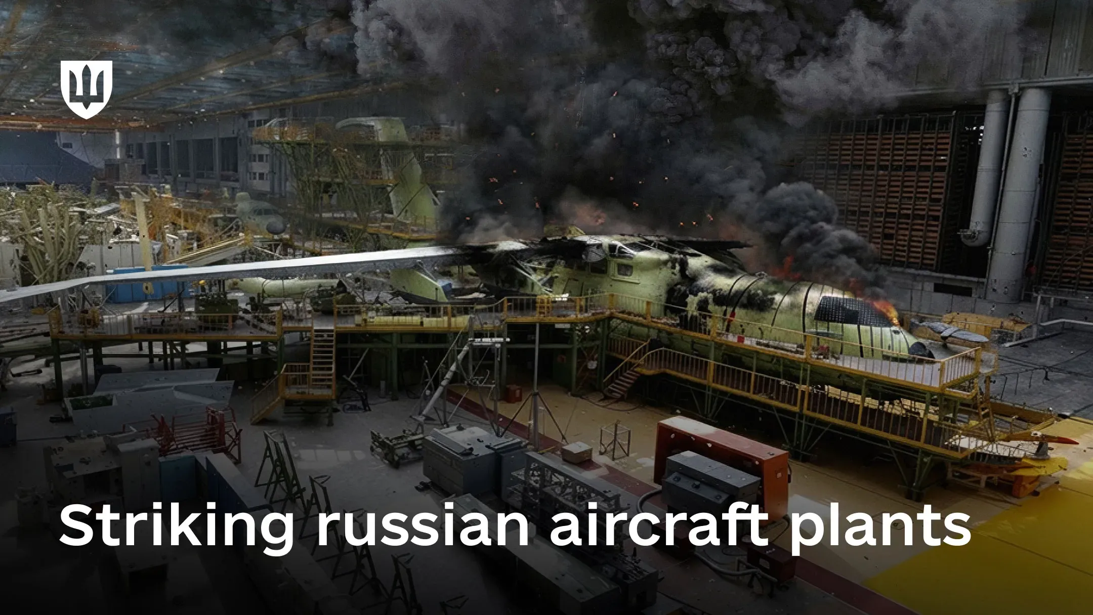 Inside an aircraft plant, a burning aircraft fuselage at the assembly stage is engulfed in flames, with the facility filled with thick black smoke.