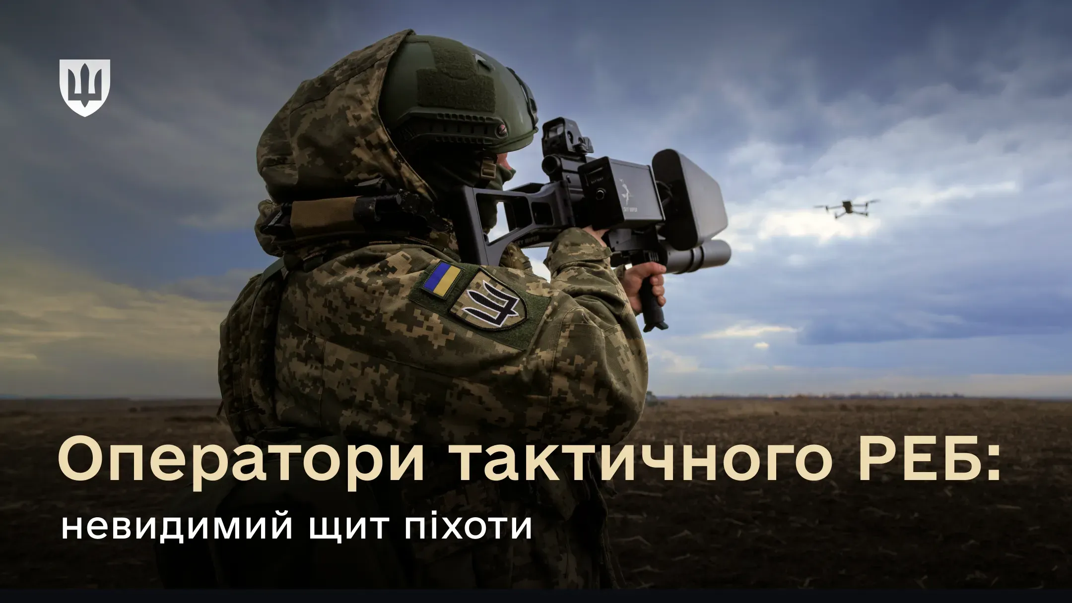 Український солдат у «пікселі» та тактичному шоломі цілиться з антидронової рушниці (комплексу РЕБ) у невеликий квадрокоптер, що завис у похмурому небі. Зйомка з-за спини військового