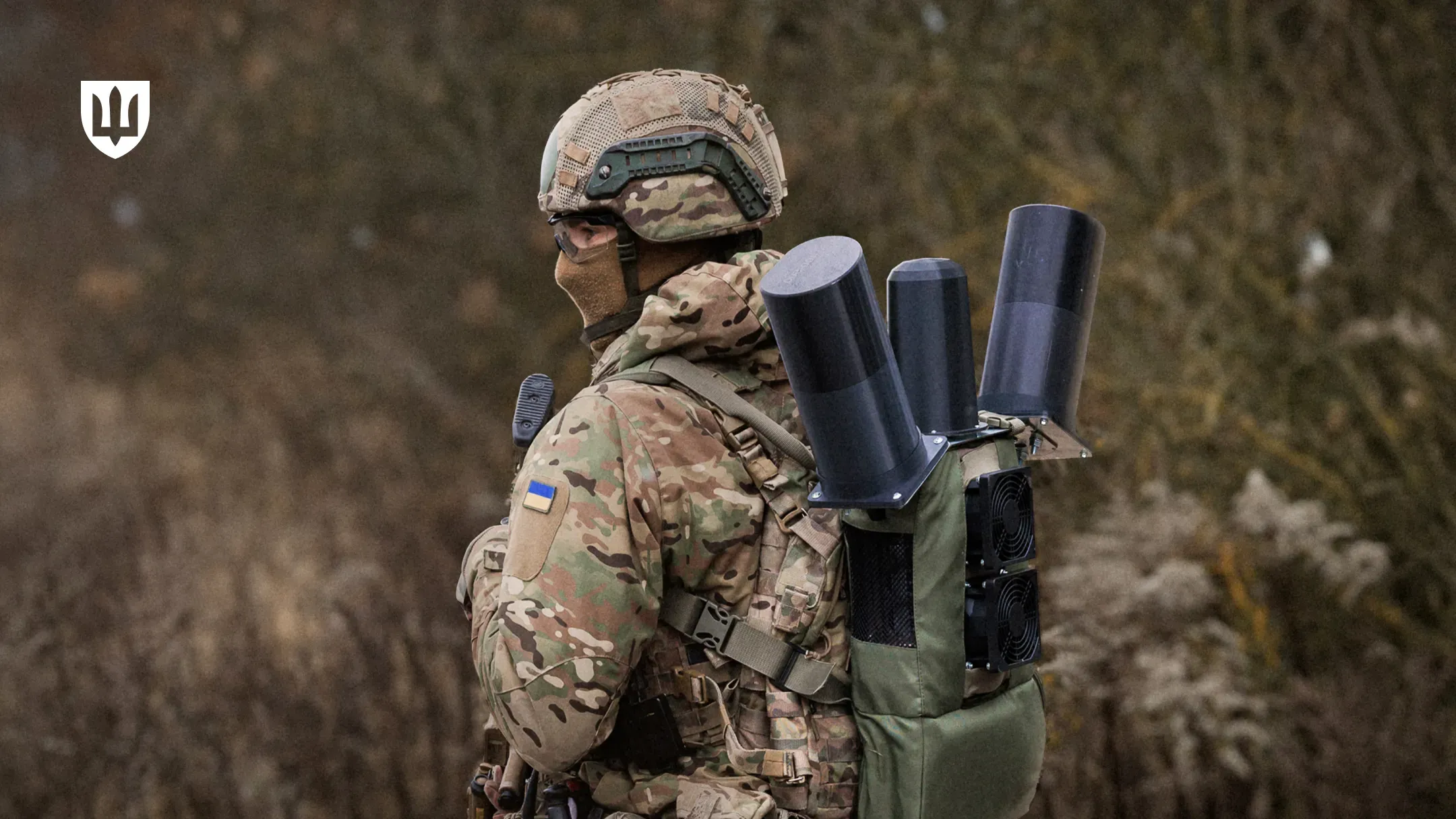Український військовий у камуфляжі Мультикам та балаклаві стоїть спиною до камери у польових умовах. На його наплічнику закріплений портативний комплекс РЕБ (окопний РЕБ) з трьома великими чорними циліндричними антенами та системою охолодження (кулерами)