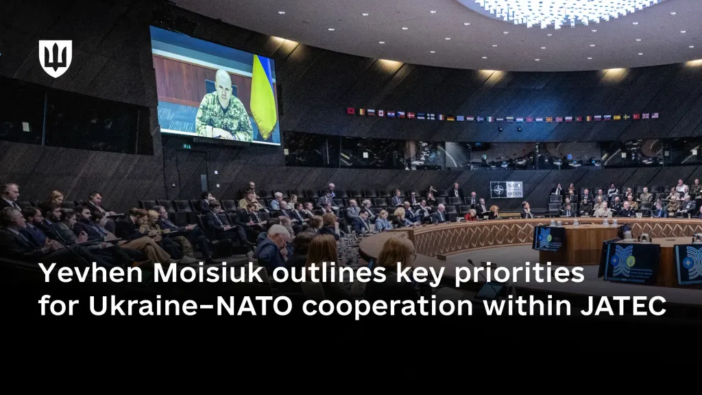 A large circular conference hall during a meeting. On a large screen, Lieutenant General Yevhen Moisiuk, Deputy Minister of Defence of Ukraine, appears in pixel-pattern uniform while addressing the participants.