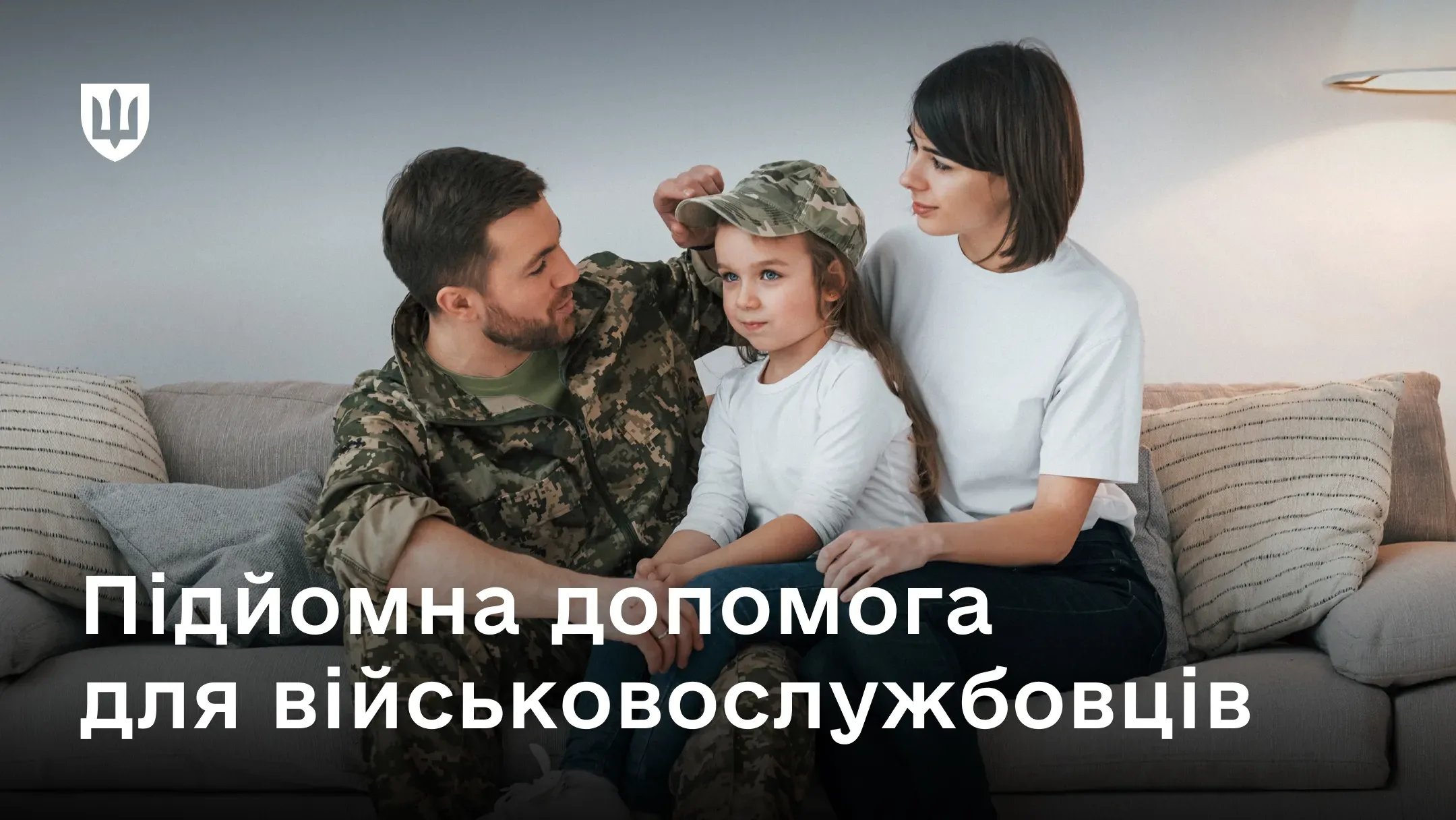 Військовослужбовець у камуфляжній формі грається з дитиною, одягаючи їй військову кепку, поруч сидить дружина