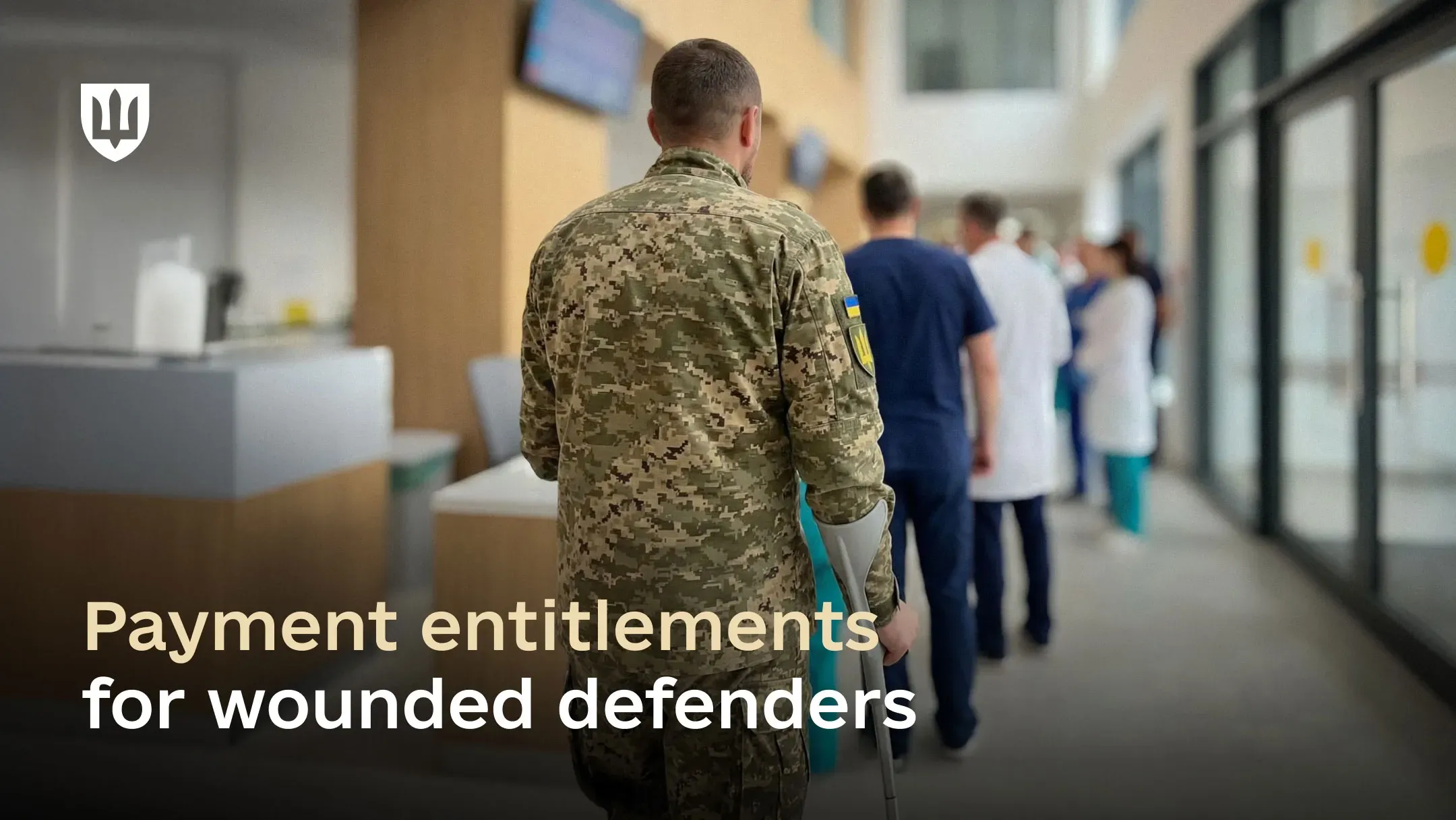 A Ukrainian service member in a pixel-pattern uniform walks through a corridor of a modern hospital using a crutch, with medical staff visible ahead.