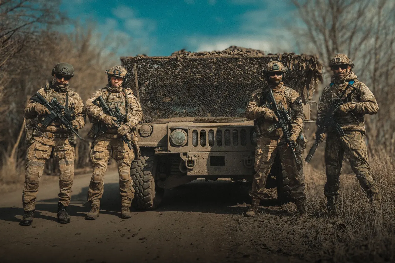 Четверо військовослужбовців у повному бойовому спорядженні та зі зброєю в руках на фоні військового позашляховика на ґрунтовій дорозі.