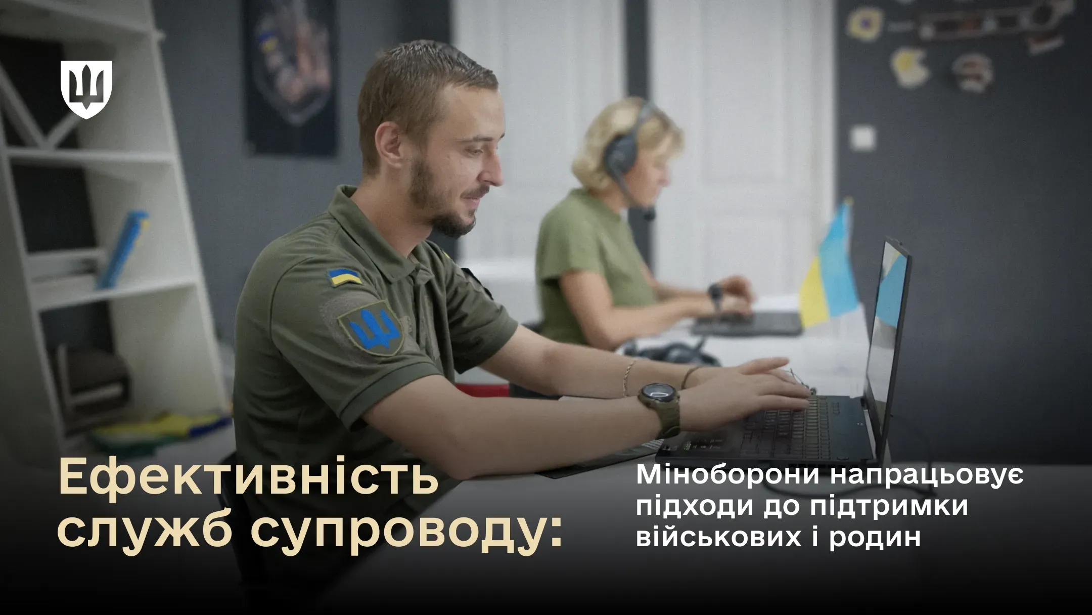 Військовослужбовець у формі ЗСУ працює за ноутбуком у офісі, на задньому плані колега з гарнітурою; на столі стоїть прапорець України.