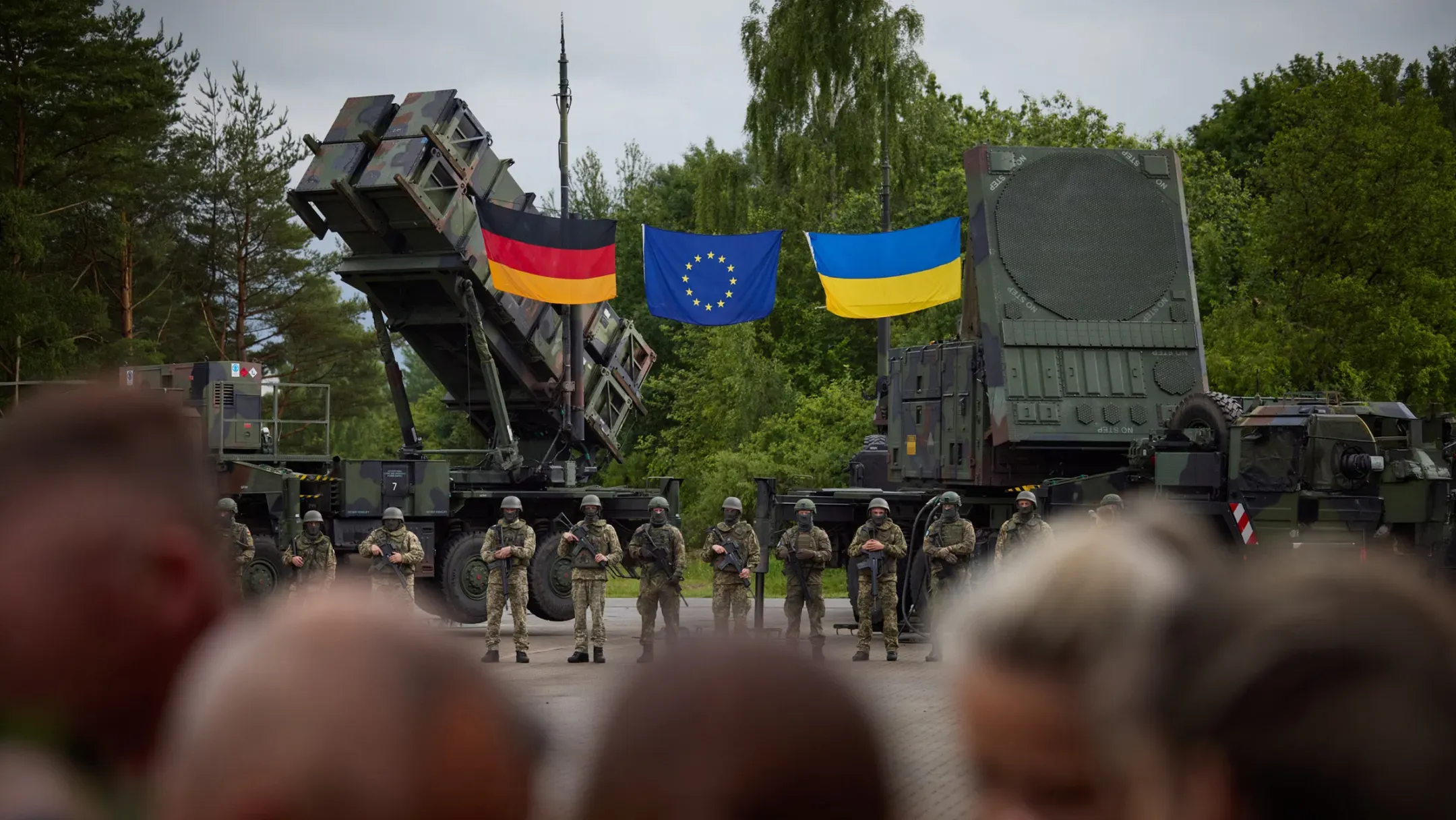 На фото військові стоять перед системами ППО Patriot на тлі дерев. Над технікою розміщені прапори Німеччини, Європейського Союзу та України